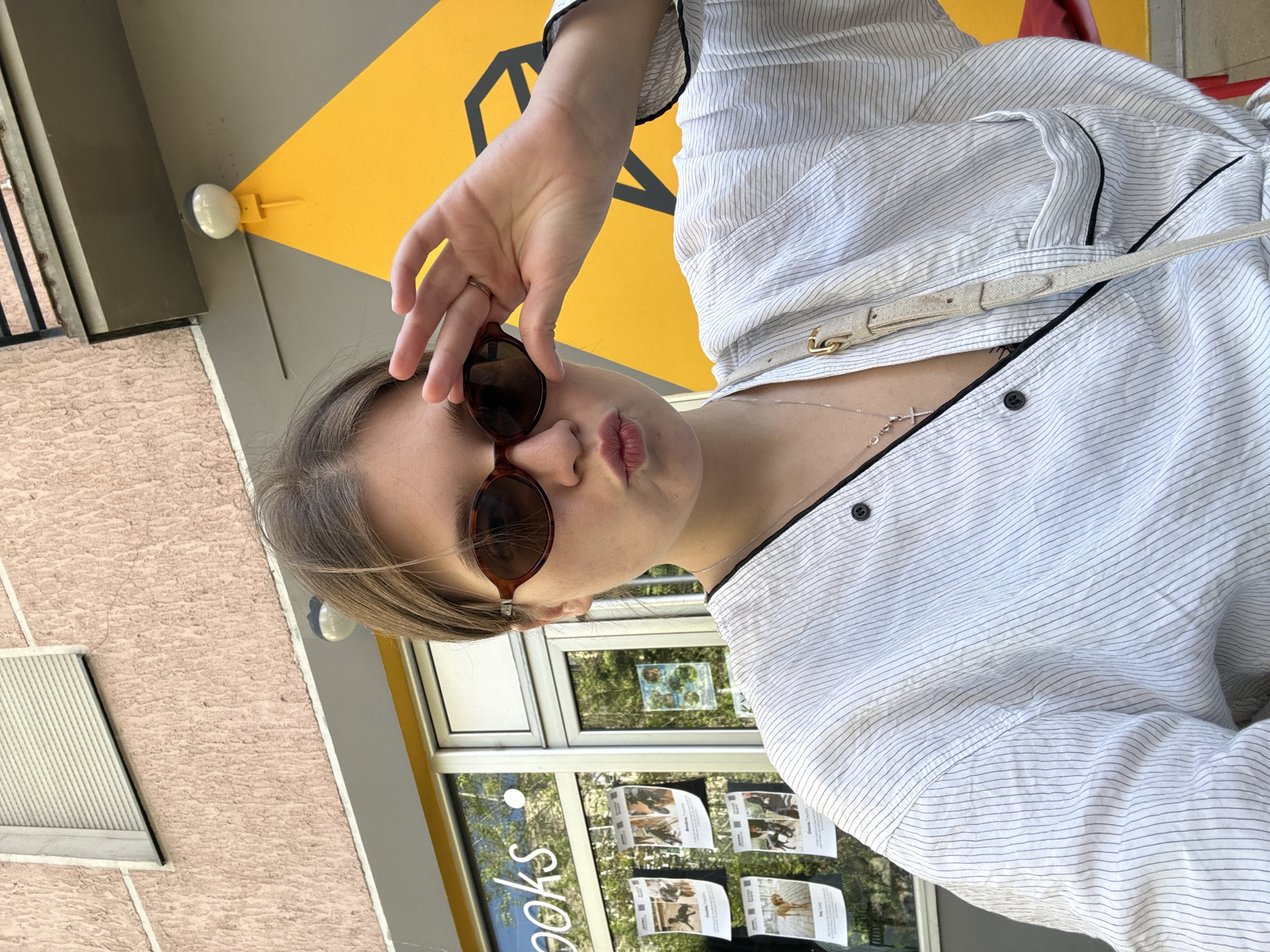 A young woman is standing outside a building in the Palilula Urban Municipality in Serbia. She is wearing a white striped shirt and sunglasses, and holding the sunglasses with her hand. There is a yellow and black sign on the building behind her. There are posters displayed on the building's window.