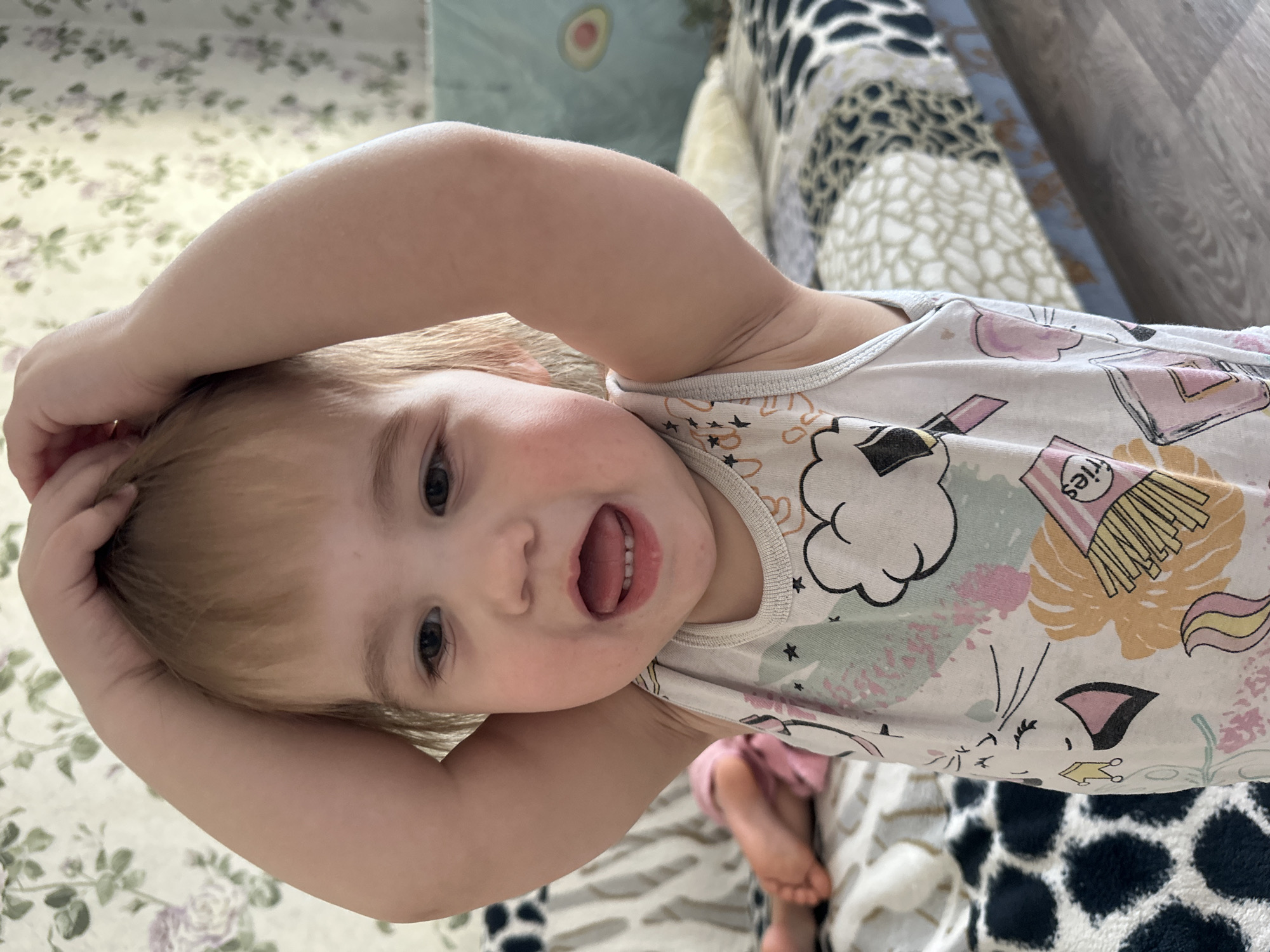 A toddler with light blonde hair is lying on their back, arms raised above their head with their hands touching. The child is wearing a white tank top with cartoon-like designs, including images of clouds, fries, and cats. The child's mouth is open, revealing their teeth. The background includes a floral pattern on the left and a multi-patterned textile on the right. The location is in Chaikovsky, Russia.