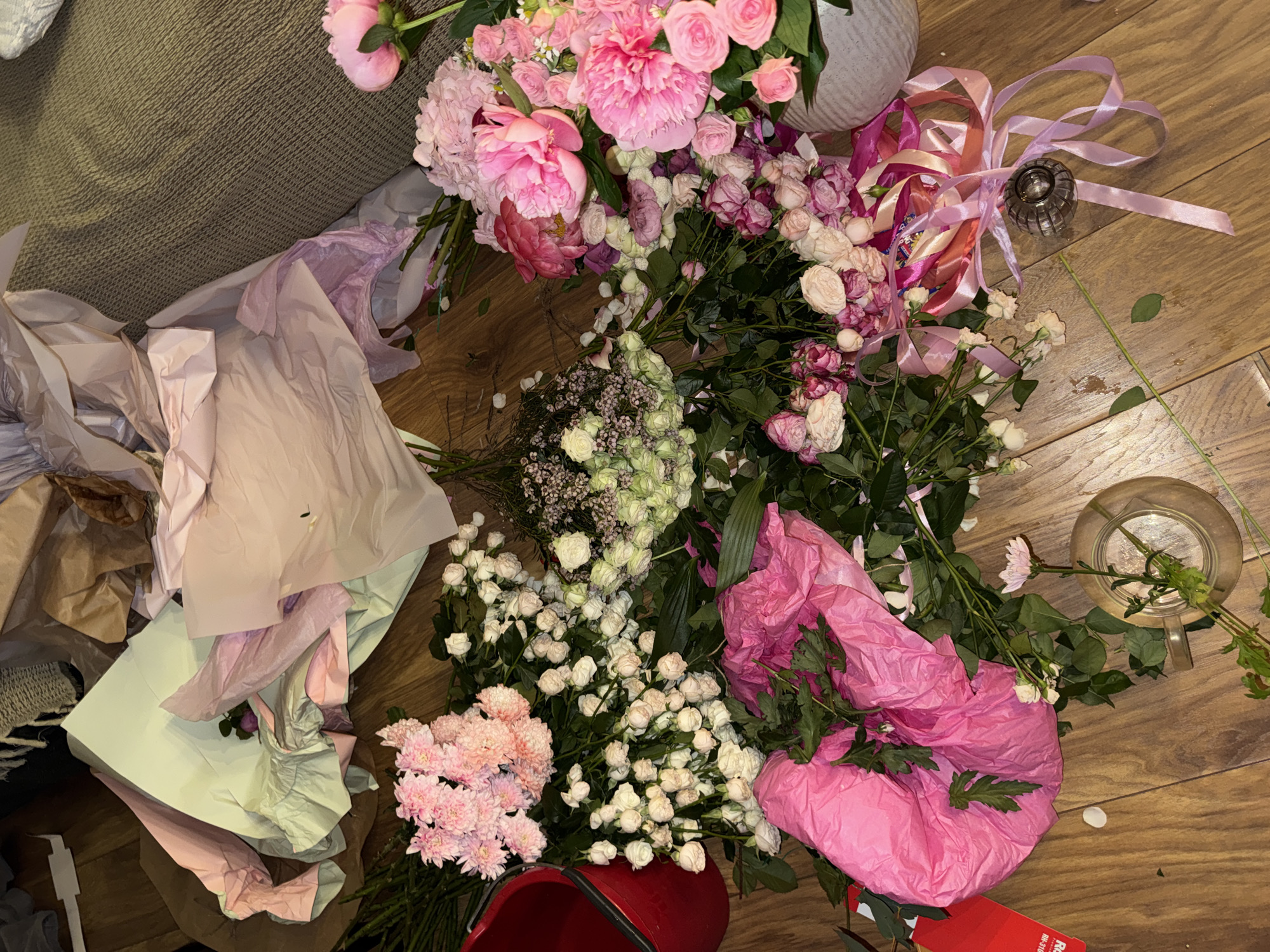 The image shows a collection of flowers and wrapping materials scattered on a wooden floor. Predominantly pink and white flowers, including roses and peonies, are spread across the floor. Some are bundled together, others are loose. Wrapping paper of varying colors (pink, white, green, brown) is also present. There are also vases and pink ribbon. The location is Unknown, Unknown. No people are visible.