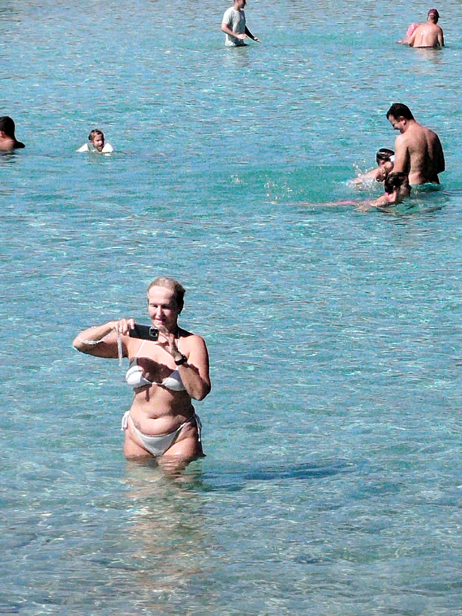 In the clear, shallow waters of Sithonia Municipal Unit, Greece, several individuals are enjoying the sea. In the foreground, a woman in a white bikini stands waist-deep, holding a smartphone to take a picture. Further out, a man with dark hair is partially submerged, facing away from the camera. To his left, another person, also in the water, is visible. In the background, a man in a white t-shirt stands in the water, looking towards the left of the frame. Another person, with their back to the camera, is visible in the upper right corner, in deeper water. The sea is a vibrant turquoise, with sunlight reflecting off its surface, creating a shimmering effect.
