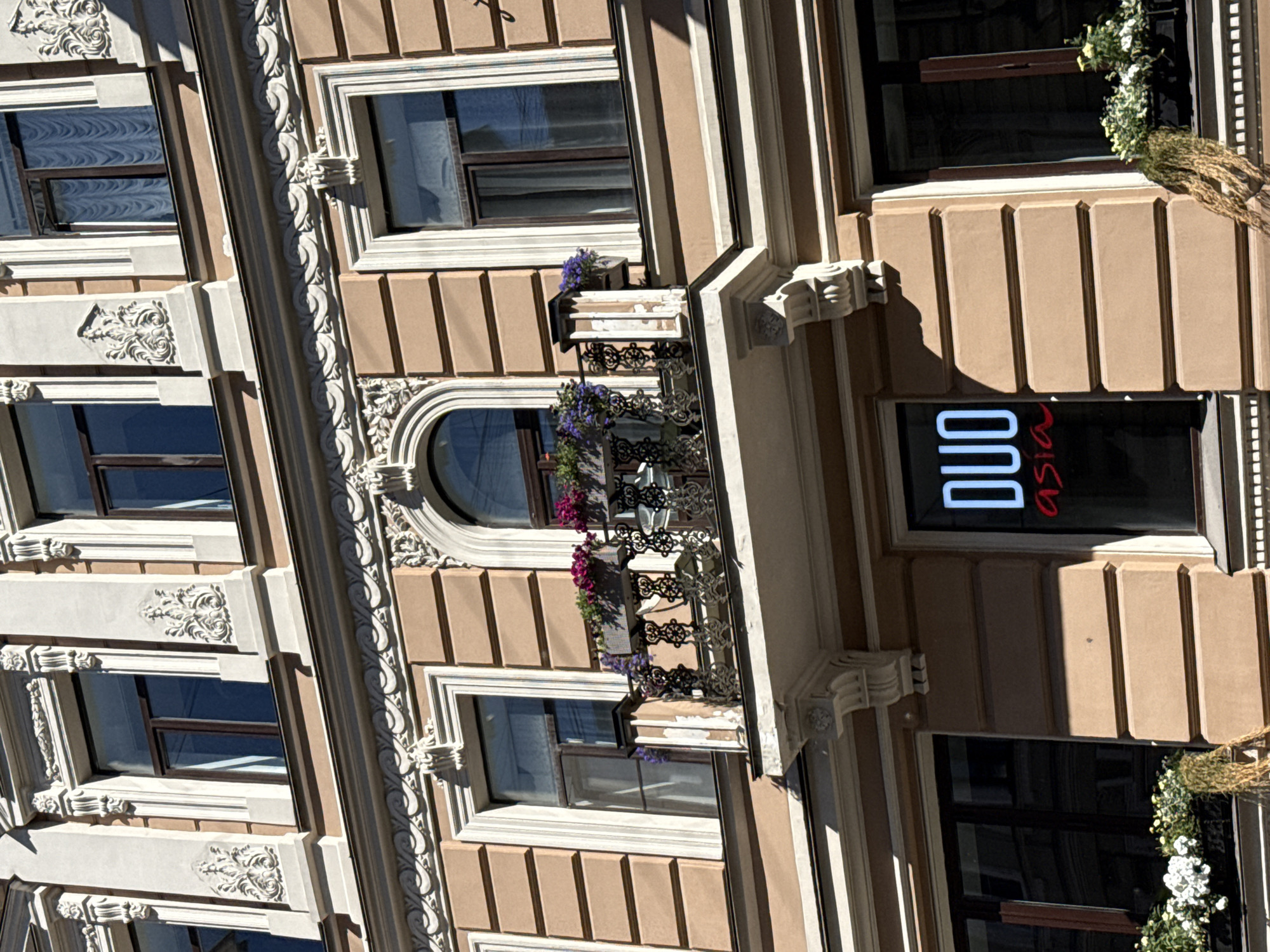 The image shows the exterior of a building with ornate architectural details, likely in Saint Petersburg, Russia. The building has a beige facade with cream-colored decorative elements around the windows and along the vertical lines of the building. There are multiple floors visible, each with several windows. Some of the windows are rectangular, while one on a lower floor is arched.
A prominent feature is a balcony with black wrought-iron railings and hanging flower baskets filled with purple and pink flowers. Above the balcony, on an upper floor, there is a window with a small box containing purple flowers.
On one of the lower floors, there is a dark storefront with illuminated text that reads "DUO Asia". The "DUO" is in white, blocky letters, and "Asia" is in red script. Above this storefront, there is a window decorated with a garland of white flowers and some dry, straw-like material. The building appears to be well-maintained and displays a classical architectural style.
The lighting suggests it is daytime, with strong sunlight casting shadows, indicating a sunny day. The overall scene evokes an urban setting with a touch of elegance due to the building's design and the floral decorations. No people are visible in the image.