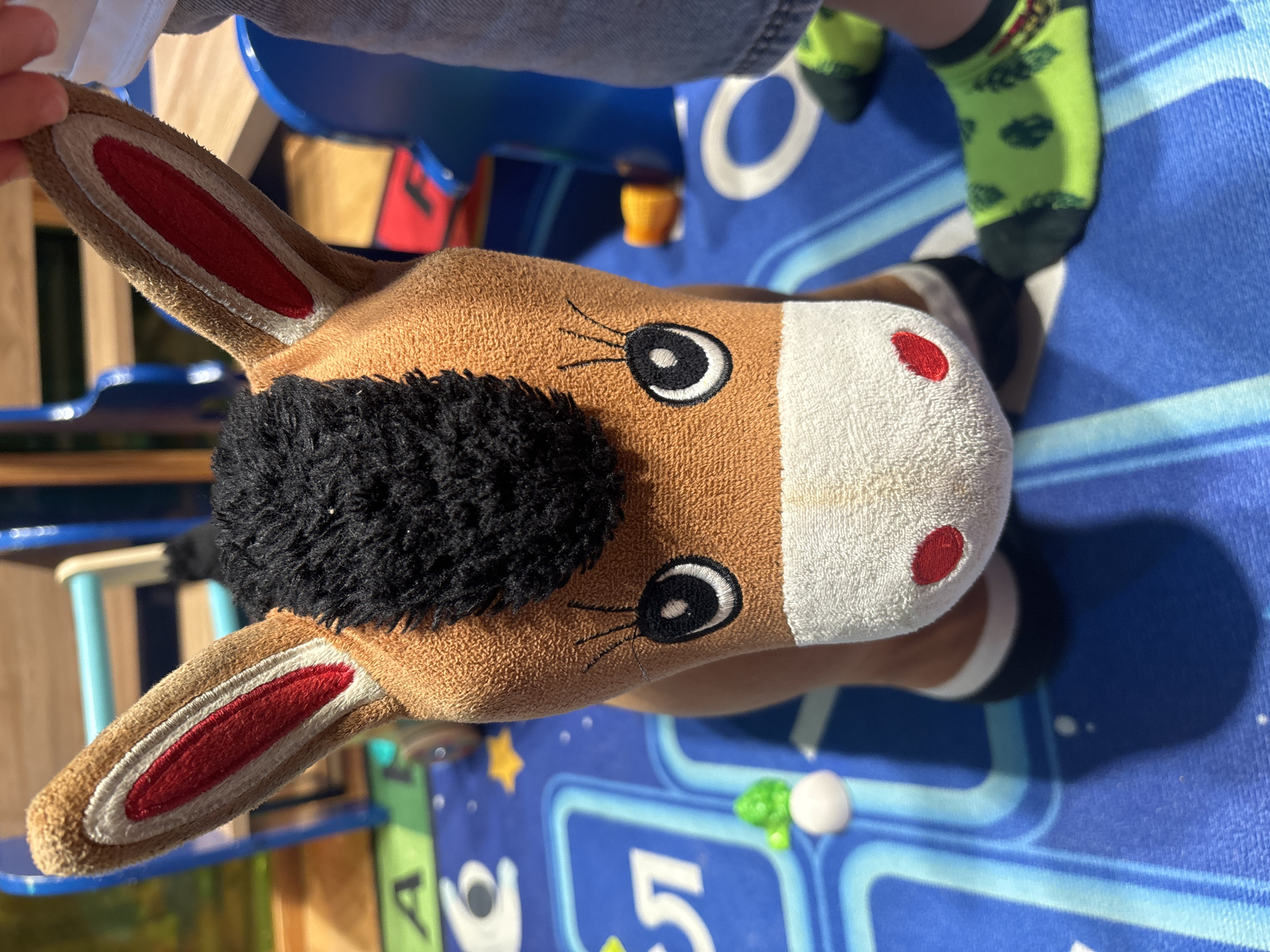 A stuffed toy horse with black mane and brown body sits on a blue mat with a playful design. The horse has red and white striped ears and embroidered eyes. A child's legs, wearing green socks with a pattern, are visible to the right of the horse, with one foot touching the toy. The background shows parts of a blue plastic structure, possibly a play area, and a wooden surface. The scene appears to be an indoor play area in Dubai, United Arab Emirates.