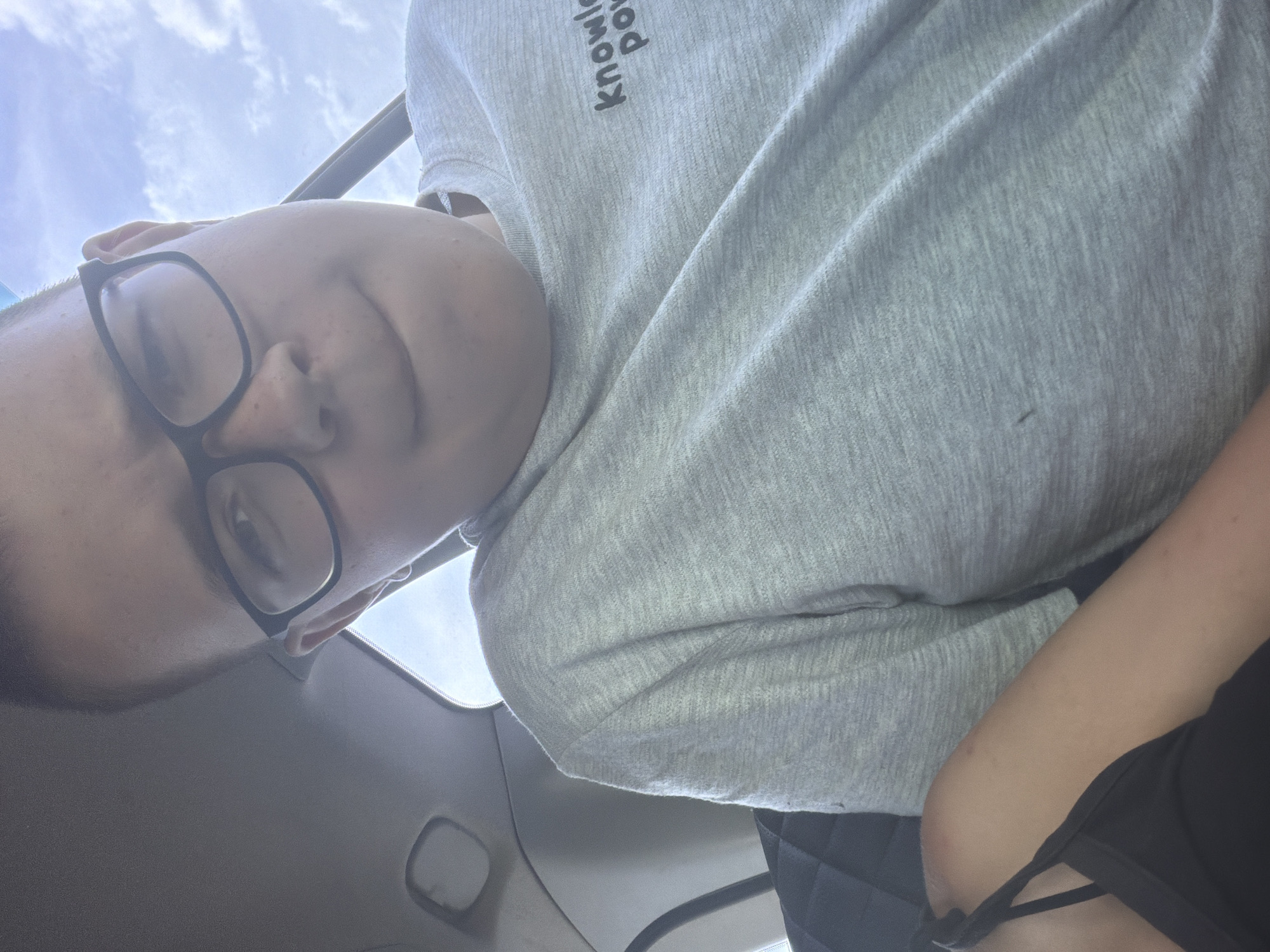 The image shows a close-up, waist-up shot of a young person wearing glasses and a light grey t-shirt with the words "knowledg pow" visible in black text. They are sitting in what appears to be a car, with a portion of a black car seat visible on the left side of the frame. The background includes the car's interior ceiling and a bright sky with clouds. The person is looking slightly down and to the right, with a neutral expression and their lips slightly parted. There is a black strap visible on their left shoulder, suggesting they are wearing a tank top or similar garment underneath the t-shirt. The image does not provide any information that would indicate it is located in Ostrovka, Russia.