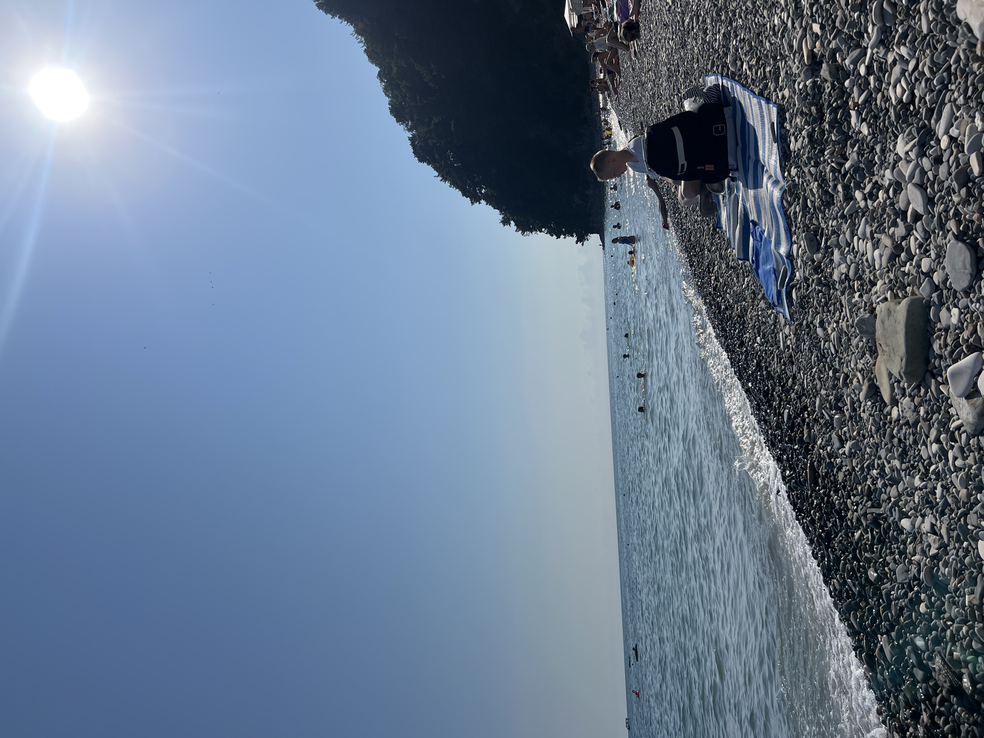 In Tuapse, Russia, on a sunny day at a pebble beach, a young boy sits on a striped mat with a black backpack beside him. The sun shines brightly in the clear blue sky, casting a warm glow over the scene. The sea is calm, with gentle waves lapping the shore. In the background, a lush green forest covers a steep hillside, adding a natural beauty to the coastal landscape. Several people are scattered along the beach and in the water, enjoying the pleasant weather. The boy appears to be relaxed, perhaps taking a break from swimming or playing in the sand. The overall atmosphere is one of leisure and enjoyment.