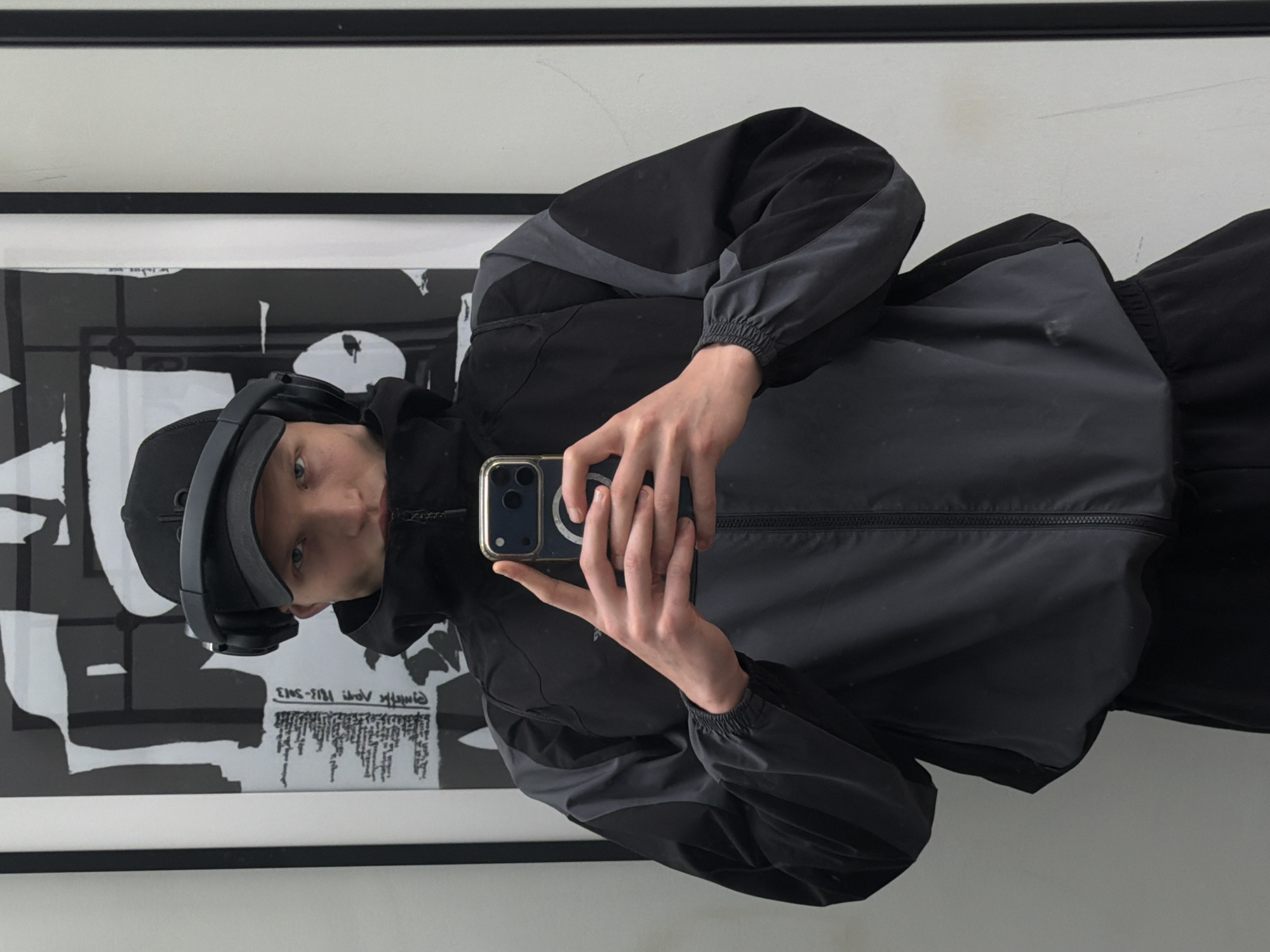 A person wearing a black baseball cap, black headphones, and a black and grey jacket is taking a selfie with a gold-cased smartphone in front of a framed black and white artwork on a white wall. The location cannot be determined from the image.