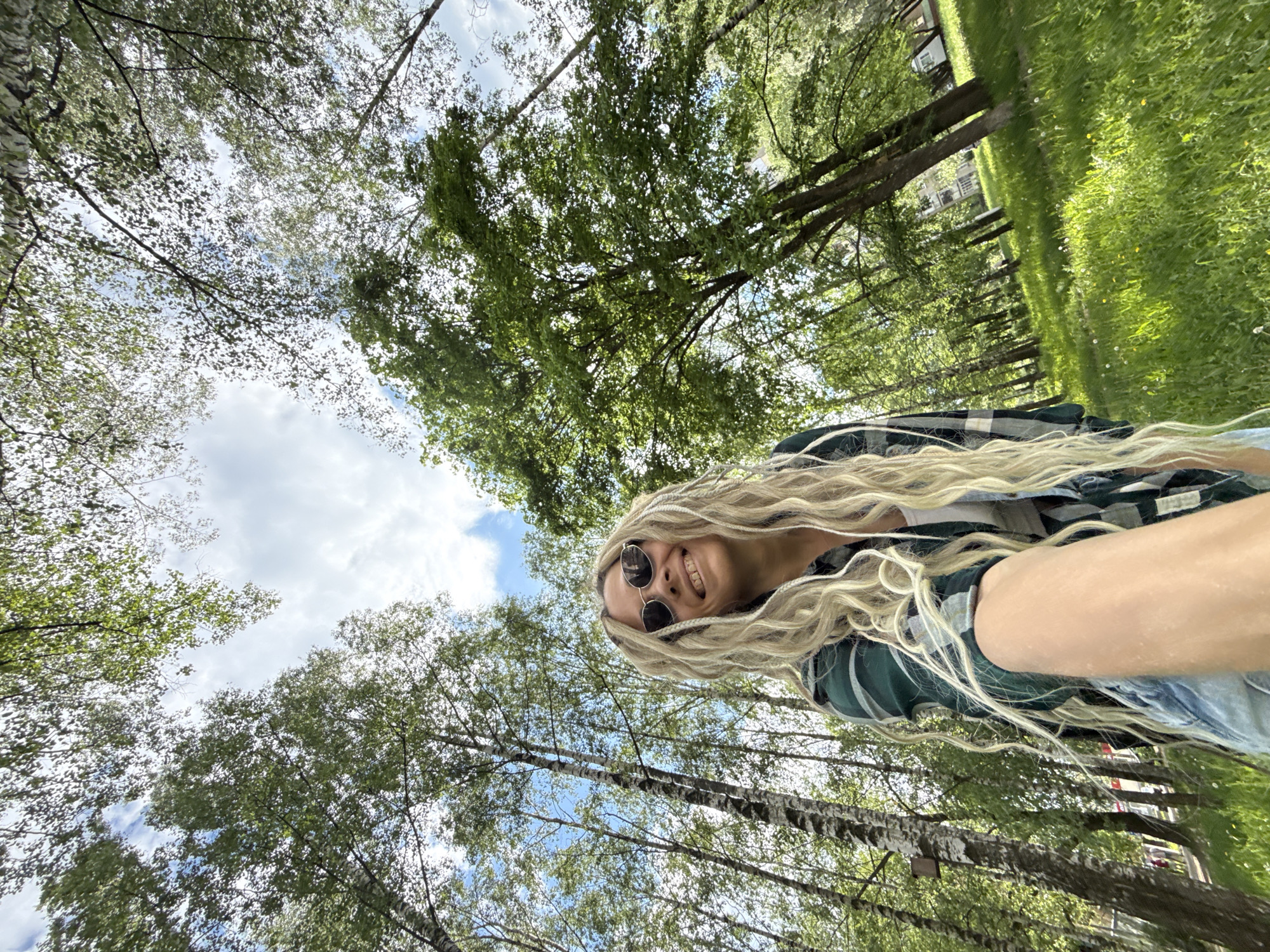 The image shows a woman in Rusanovo, Russia, from a low-angle perspective. She has long, wavy, blonde hair and is wearing sunglasses and a green and gray plaid shirt. She is smiling. The background consists of trees and a cloudy sky, suggesting a park or wooded area. Some green grass and a building are visible in the distance.