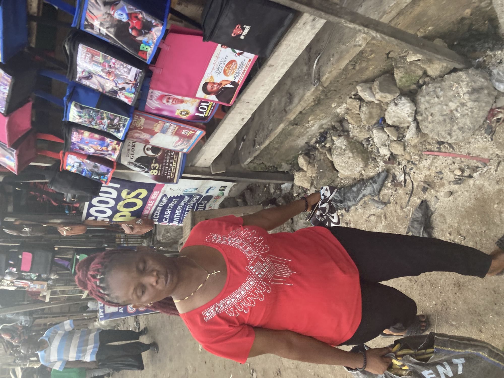 Here's a detailed description of the image:
**Setting:**
* The image appears to be taken in a bustling outdoor marketplace or street-side vending area in Ura Nta Umuarandu, Nigeria. The ground is a mix of concrete and dirt, scattered with debris.
**People:**
* The main subject is a woman wearing a red shirt with silver embellishments, black pants, and sandals. She has dark hair styled in red-tinged braids. She is looking down.
* Part of a man wearing a blue and white striped shirt and black pants is visible in the lower left.
**Objects:**
* Behind the woman, a wooden shelf displays various items:
* Lunchboxes or bags featuring cartoon characters and other designs.
* Posters or banners with images of people (likely religious figures or local personalities). Some have text and dates, including "83" and "1940-2023." The words "CHIEF ROBINSON ONYEMACHI OKI" are also visible on one. Other posters read “Lou Oparanwata” and “Forever Innocenta Nnenna.”
* Advertising posters, reading “Goody POS” and “Available Here,” advertising banking transactions are displayed.
* The woman is holding a bag, with the word “ENT” partially visible.
**Actions and Interactions:**
* The woman appears to be either looking into a bag or engaging with something at her feet.
**Overall Impression:**
The image shows a typical scene of street commerce in a Nigerian town, likely a small business owner tending to her wares or engaging with a customer.