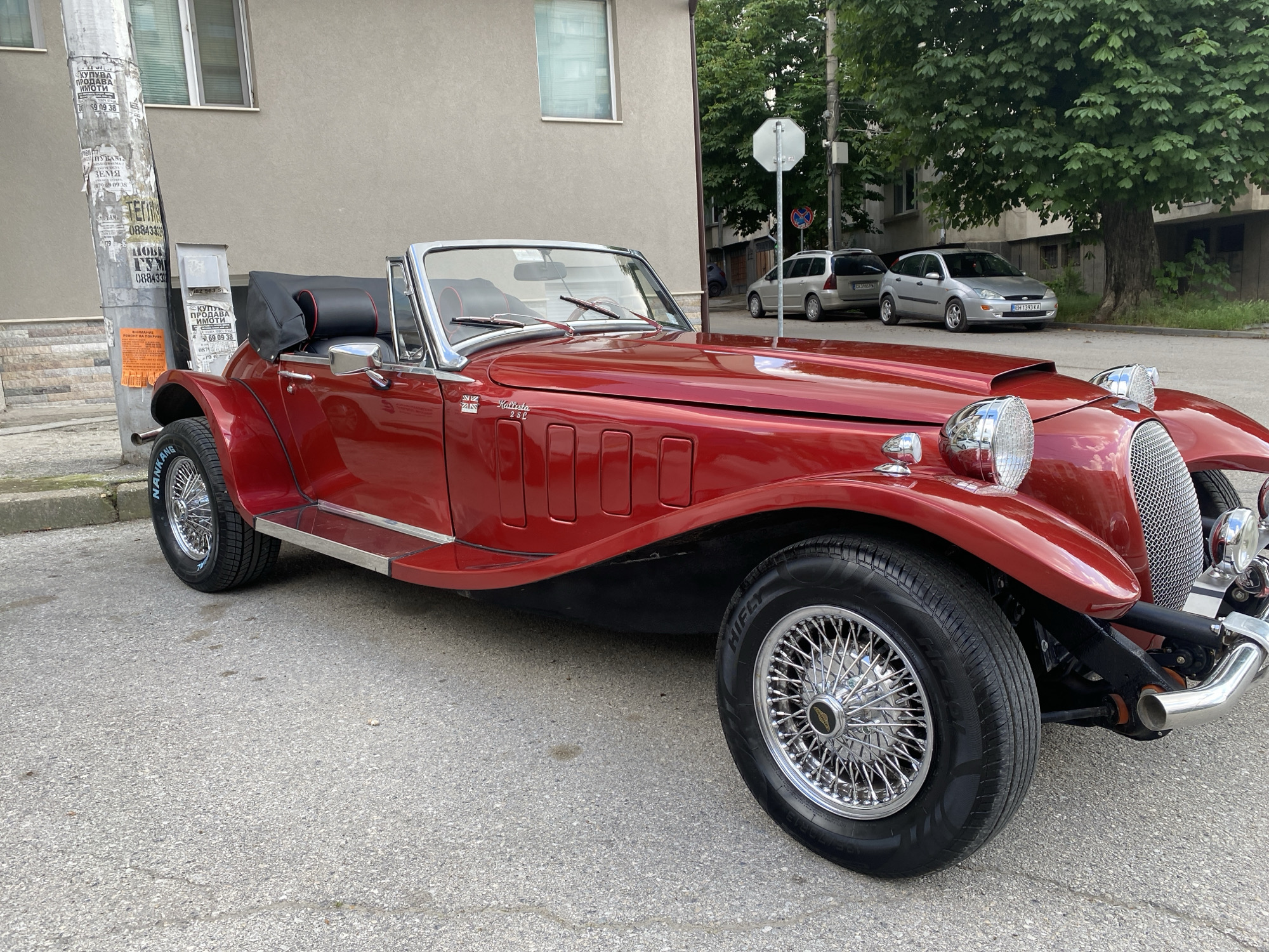 A red convertible car, a "Kallista 2.8L", is parked on an asphalt road next to a utility pole covered in posters. The car has chrome wire wheels and a black interior. A building with windows is visible in the background. Two parked cars, a silver Ford Focus ("EH 1393 KA") and a gray station wagon, are visible in the distance, along with a stop sign. The location is in the city of Pleven, Bulgaria. The word "NANKANG" is visible on the tire.