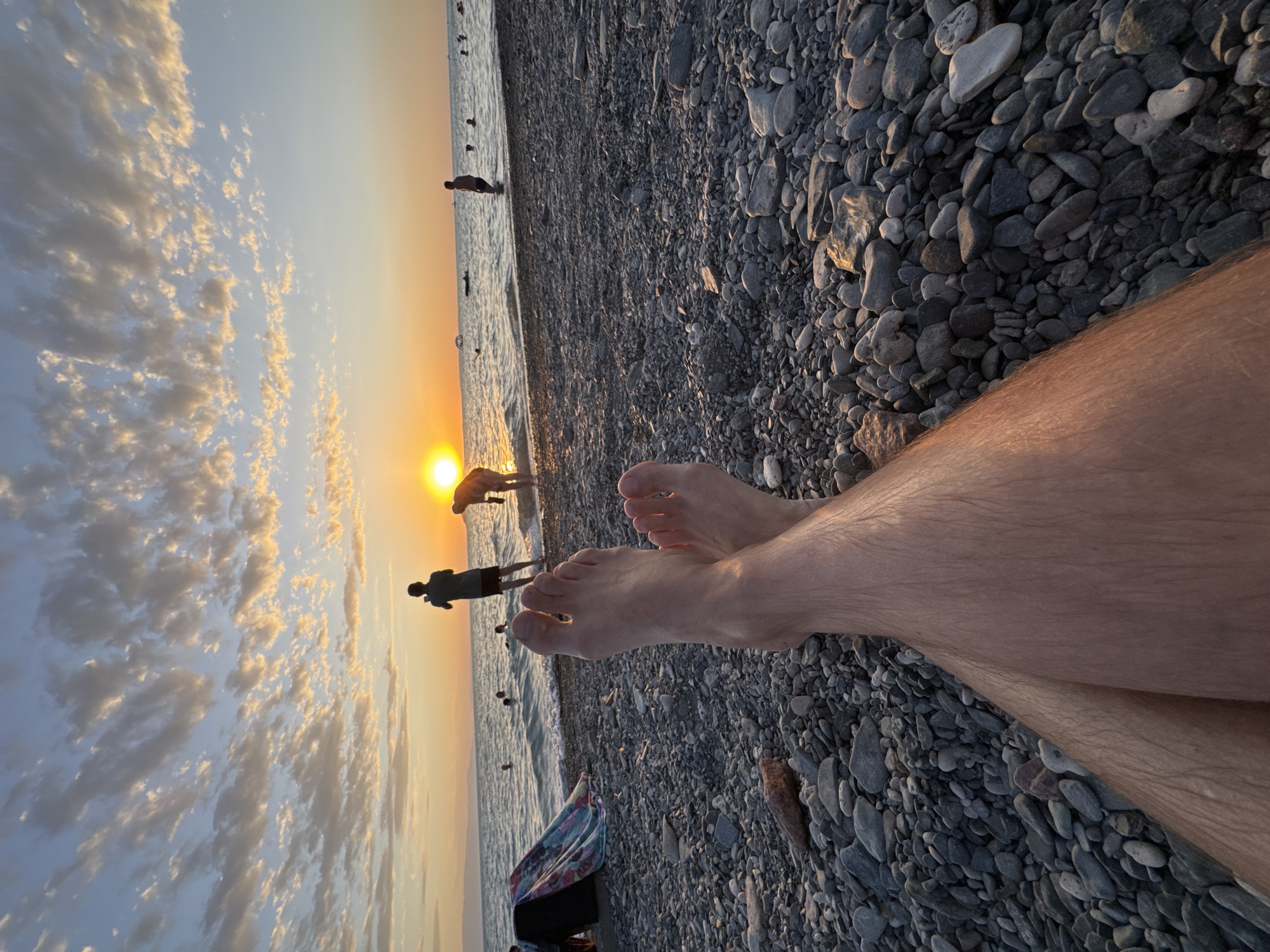 The image shows a first-person perspective of someone sitting on a pebble beach, with their legs and feet prominently in the foreground. The legs are crossed and appear to be hairy. The beach is covered in dark gray and light-colored pebbles. In the background, the sun is setting over the sea, casting a warm orange glow on the sky and clouds. Several people are visible in the water and on the shoreline. Some are wading in the shallow water, while others are standing on the beach. One person in the distance appears to be taking a photo with a mobile phone. The overall scene depicts a serene beach setting during sunset. The location is identified as the city of Zharbulak, Kazakhstan.