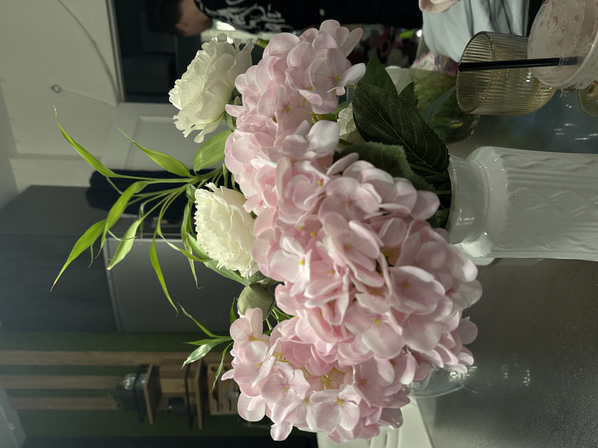 The image shows a bouquet of white and pink hydrangeas with green foliage in a white vase, sitting on a reflective surface. In the background, partially visible, are a person wearing a patterned shirt, some glasses, and a drinking glass with a straw. The environment appears to be indoors, possibly a room within a residence or a commercial establishment, as indicated by the presence of furniture or decor. The image provides no verifiable information to associate the location to the city of Селекционер-2 Снт, Russia.