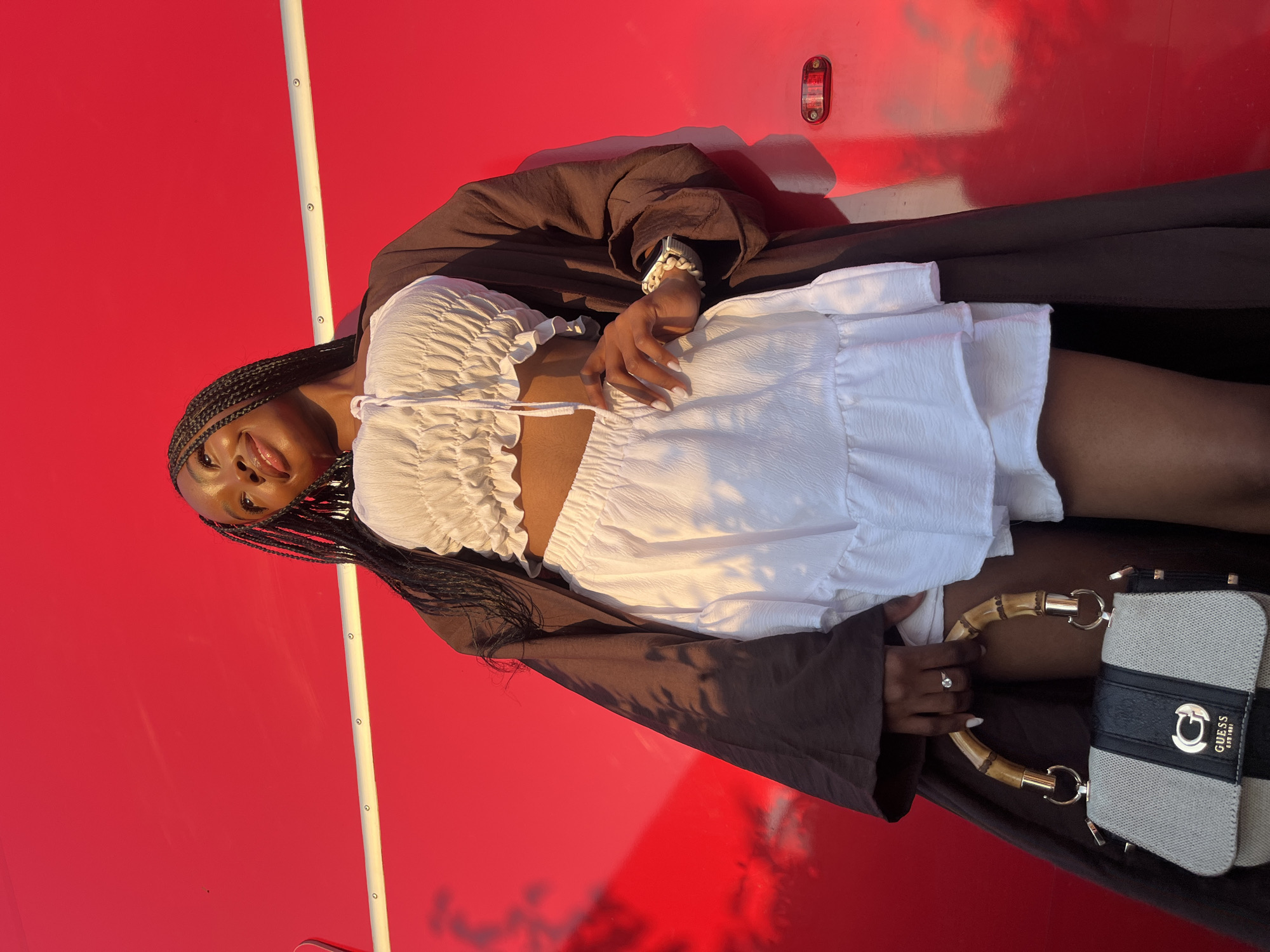 A young woman with braided hair stands against a vibrant red wall. She is wearing a white crop top and a white ruffled skirt, covered by a dark brown open coat. She holds a gray and black purse with a bamboo handle. The woman is looking up and to the right, with a slight smile. The red wall has a horizontal white bar with circular fasteners running across it. Shadows of foliage are cast on the wall to the left. The image does not contain any elements that indicate the city of Kazan, Russia.