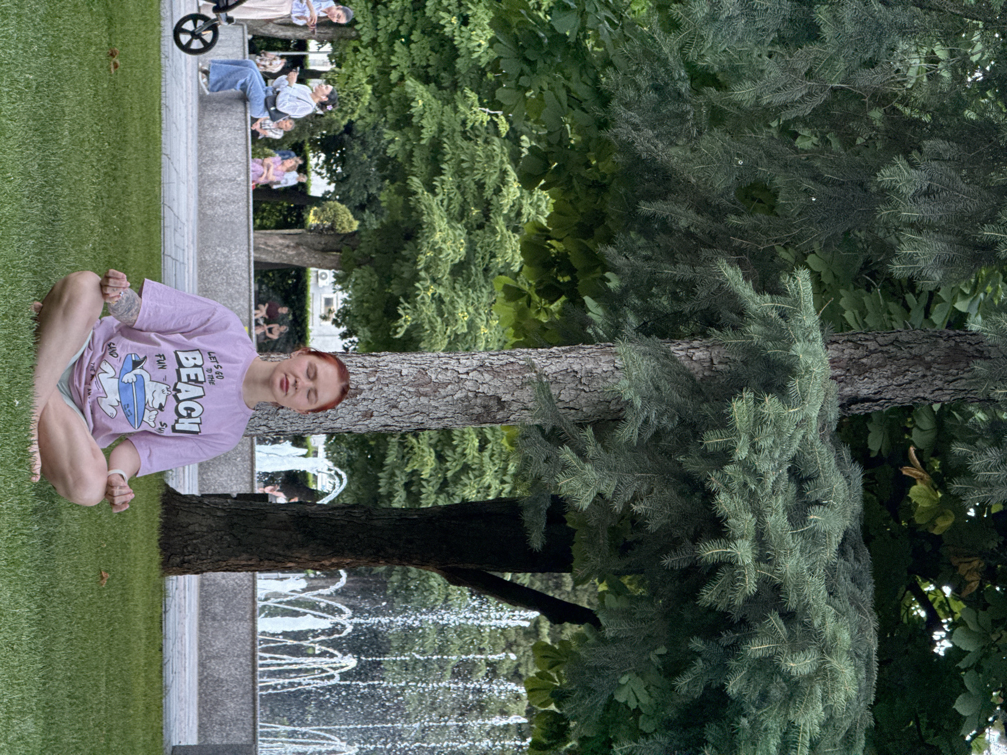 This image shows a young woman with red hair sitting cross-legged on a grassy lawn in a park. She is wearing a light purple t-shirt with a cartoon character and the words "LET'S GO BEACH FUN" printed on it, and light blue shorts. Her eyes are closed, and she appears to be meditating or relaxing. In the background, several people are seated on a raised stone platform, some looking at their phones, and one person is pushing a stroller. Behind them, trees and foliage create a lush, green environment. A fountain with water sprays is visible in the lower right corner of the image, suggesting it is a warm day. The image is oriented vertically, with the subjects appearing sideways. The overall atmosphere is peaceful and serene.
