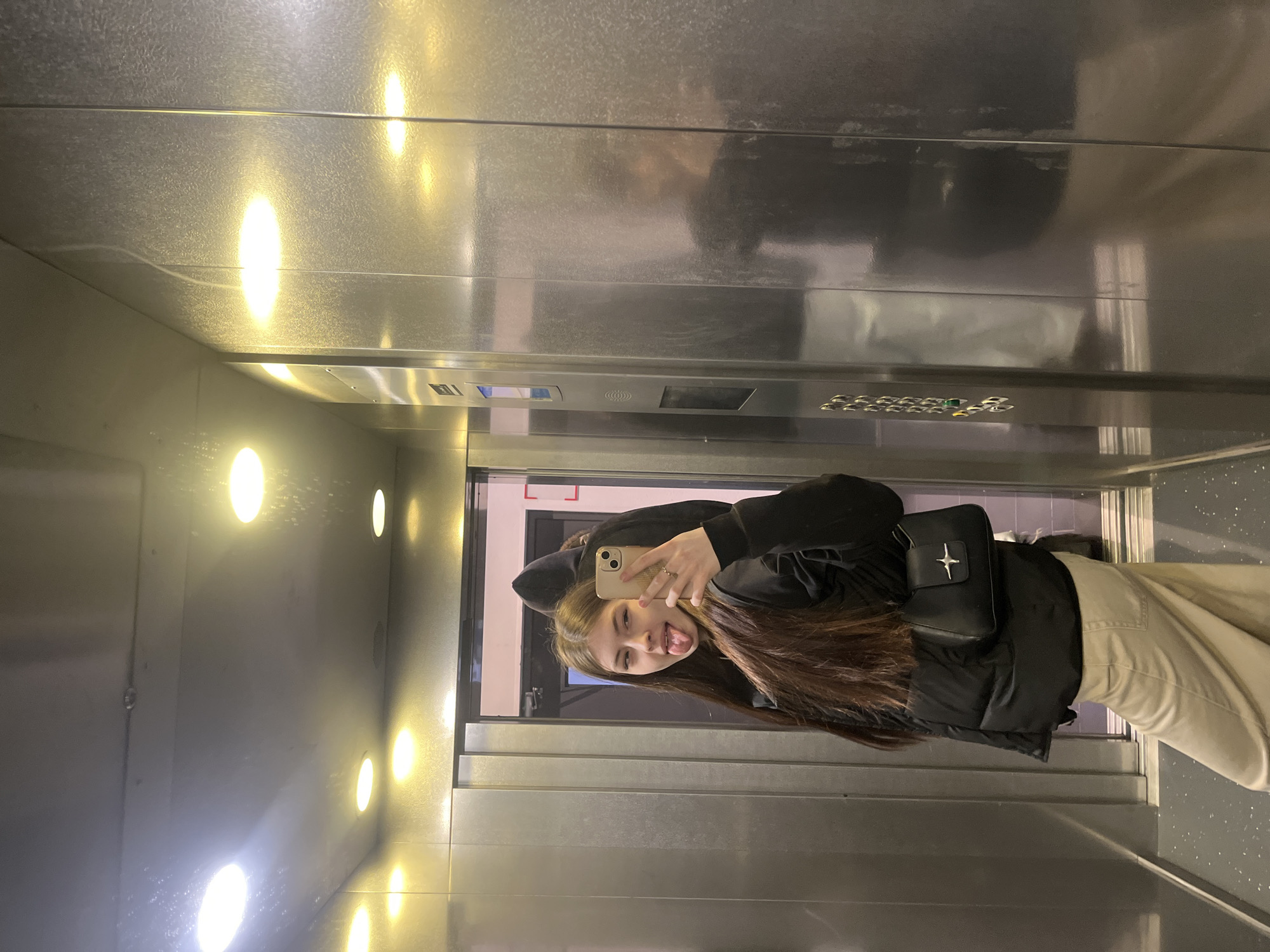 A young woman in Saint Petersburg, Russia, is taking a selfie in a mirrored elevator. She is wearing a black hooded sweatshirt, a black puffer vest, beige pants, and a black purse with a white star-like logo. She is holding an iPhone and sticking her tongue out. The elevator is stainless steel with circular lights. The elevator door is open, revealing a gray hallway and a red-framed doorway.