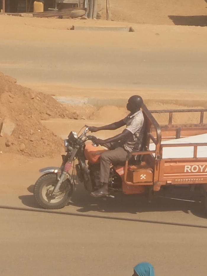 An adult male operates an orange motor tricycle, branded "ROYAL," on a light-colored, possibly unpaved road in Akwanga, Nigeria. The male, dark-skinned and with short dark hair, wears a patterned short-sleeved shirt, dark trousers, and dark sandals. His hands are positioned on the handlebars as if driving. The tricycle features a cargo bed at the rear, covered by a white tarp or sheet. An orange box with a white 'X' symbol is affixed to the side of the cargo bed. To the left, a large pile of reddish-brown earth and rocks is visible, suggesting construction or excavation. The background features distant structures and a yellow container on slightly elevated ground. The scene is illuminated by strong daylight. A portion of a person's head, covered by a light blue headscarf, is visible in the lower right foreground.