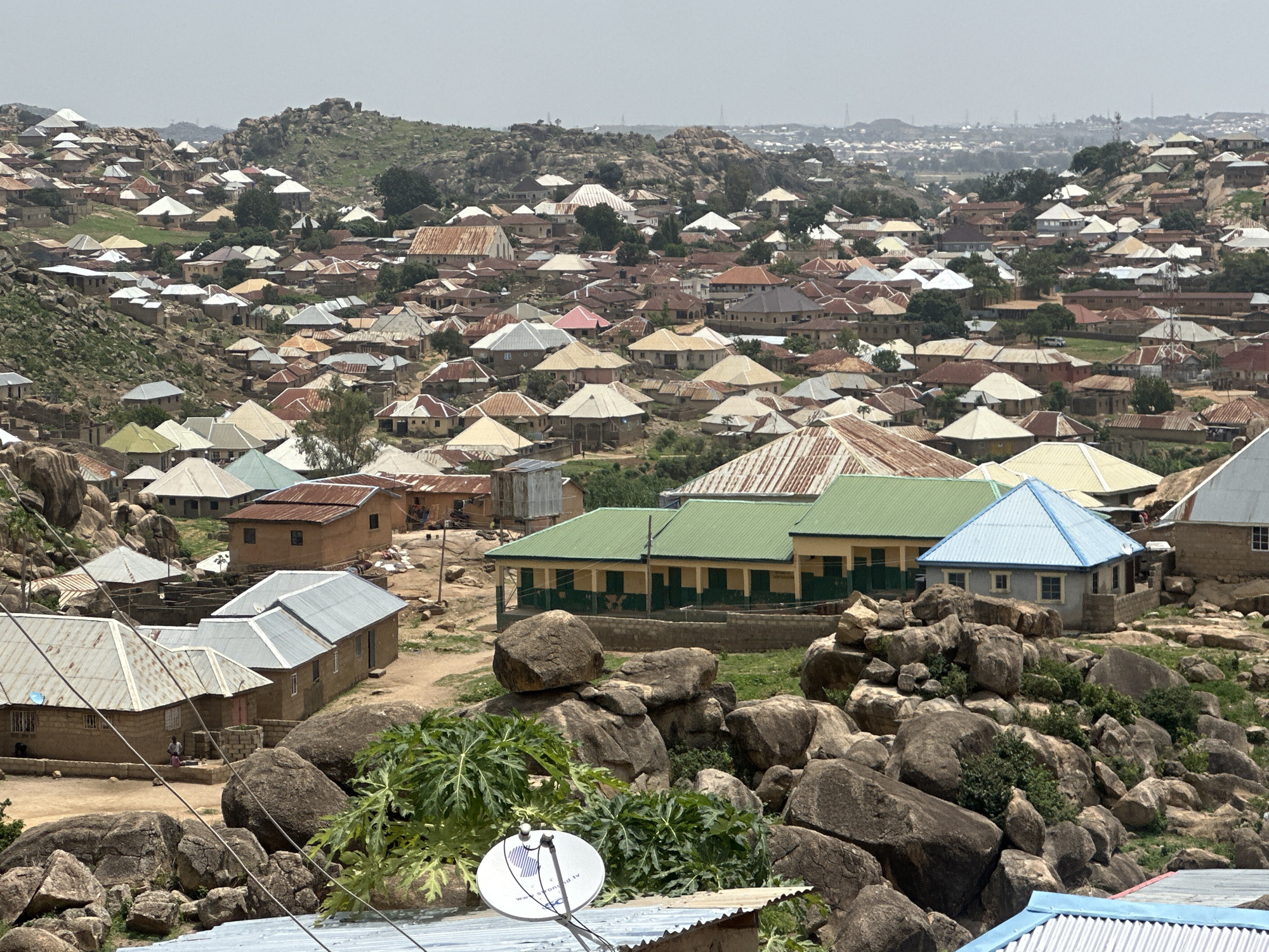 An elevated view showcases a dense residential area of Jos, Nigeria. Houses with predominantly metal roofs, painted in varying shades of brown, gray, green, and blue, cluster together over a rocky terrain. The houses are compact, with single-story structures. A building with green trim and a pale yellow façade stands prominently among the others. Boulders and sparse vegetation are scattered throughout the landscape. In the foreground, a satellite dish sits atop a corrugated metal roof. The sky is overcast. The urban landscape extends into the distant horizon, punctuated by hills and buildings. No people are clearly visible.
