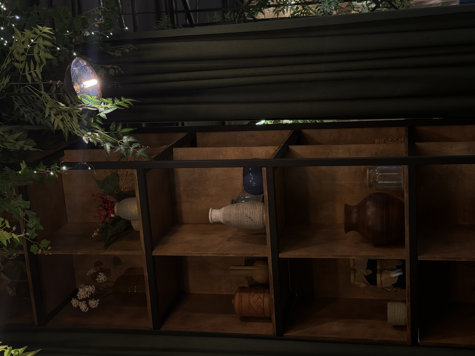The image depicts a wooden shelf unit with various decorative items. The shelves are dark wood with a black metal frame. On the shelves are vases, decorative flowers, and other small objects. The lighting is dim, with a spotlight illuminating the top left corner of the shelf unit where greenery is present. A dark, horizontal structure is above the shelf, potentially a roof or covering. The image does not show any people, actions, or interactions. There is no information about the location in Izhevsk, Russia, in the image.