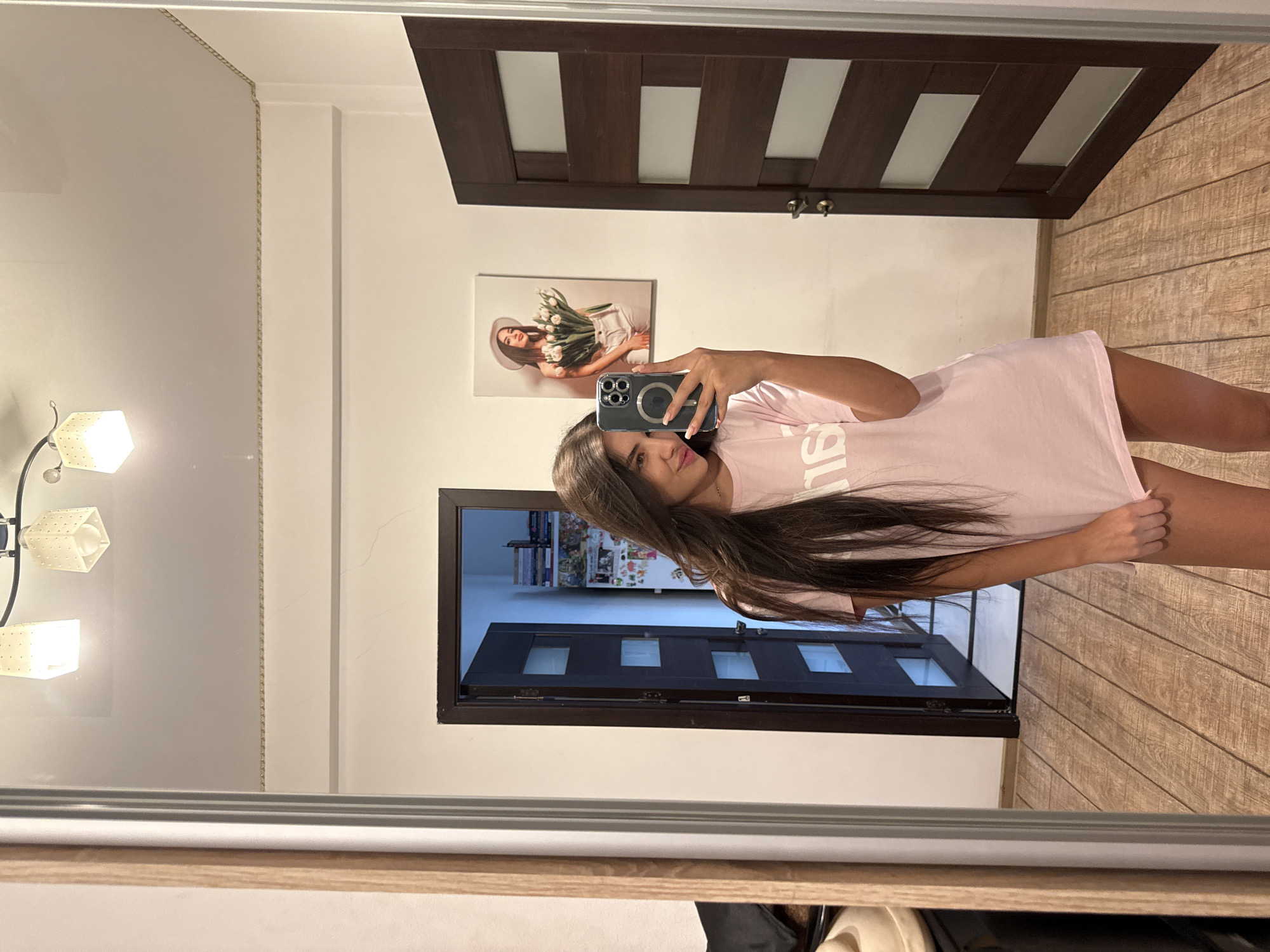 A young woman with long, dark hair is taking a mirror selfie in what appears to be a residential hallway or entryway in Chișinău, Moldova. She is wearing a pink oversized t-shirt and is holding a smartphone in her right hand to capture the reflection. Behind her, a dark wooden door with glass panels is ajar, revealing a glimpse of another room. To her left, a large mirror frames the scene, reflecting the woman, the doorway, and part of a wooden floor. On the wall to her right, a framed picture of another young woman holding a bouquet of white flowers is visible. The room is lit by overhead lights, and the walls are painted a light color. The floor is made of light-colored wooden planks.