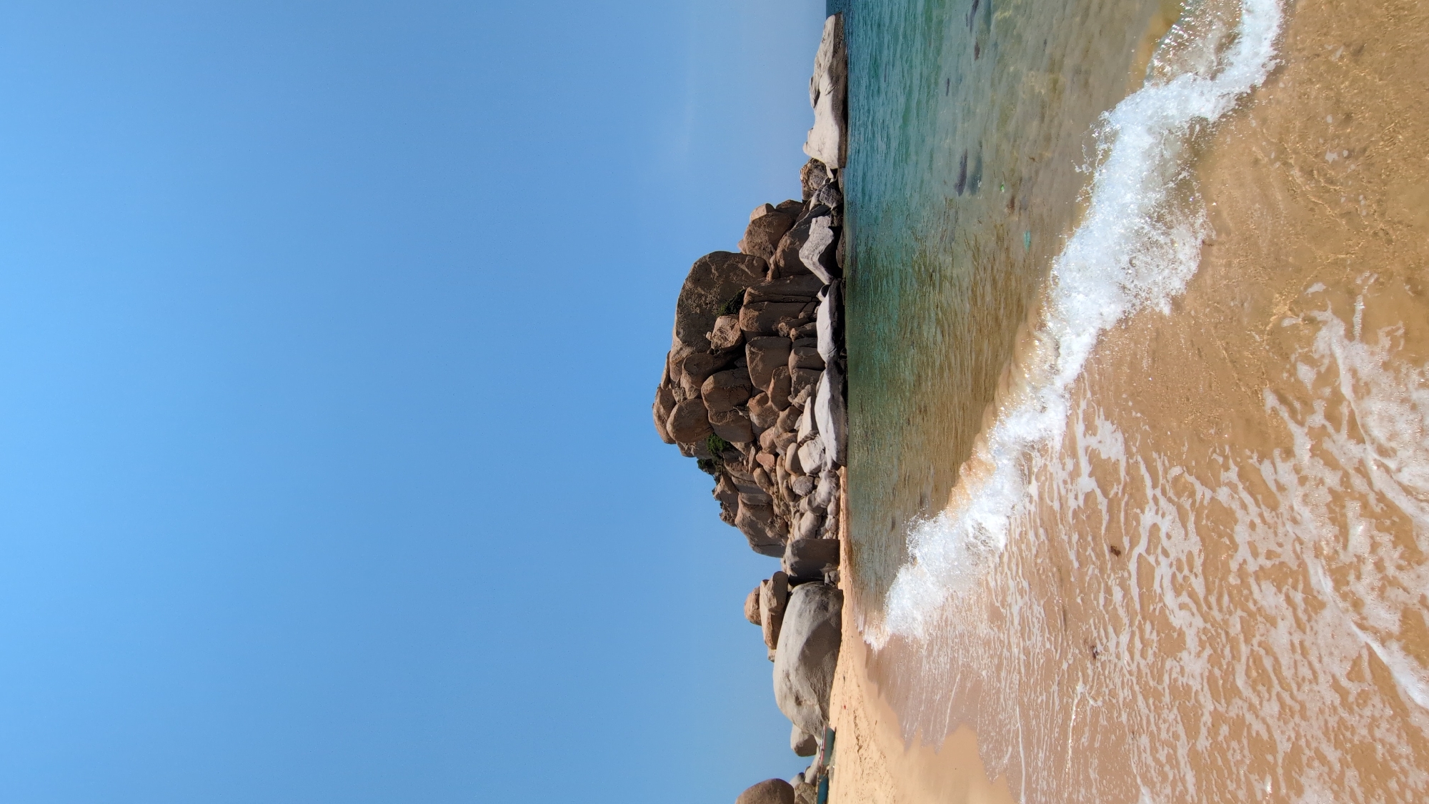 A landscape-oriented photograph captures a beach scene in Phuoc Dinh, Vietnam. The composition features a clear blue sky that dominates the left side of the frame. To the right, a rock formation rises from the beach. The beach itself transitions into turquoise water, with white waves crashing onto the sandy shore. The color palette consists primarily of blues, browns, and tans. The photograph is taken from a slightly elevated perspective, providing a panoramic view of the beach and coastline.