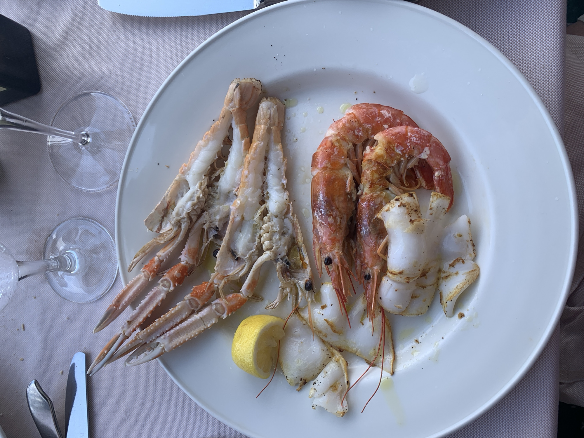 A white plate of seafood rests on a light pink tablecloth. The plate contains langoustines, shrimp, and what appears to be calamari. A wedge of lemon is also on the plate, along with a small amount of oil and black pepper. Two wine glasses are visible in the background, and cutlery is present on either side of the plate. The scene appears to be an outdoor dining setting, possibly a restaurant in Pozzarello, Italy, given the presence of seafood and wine glasses. The lighting suggests it is daytime.