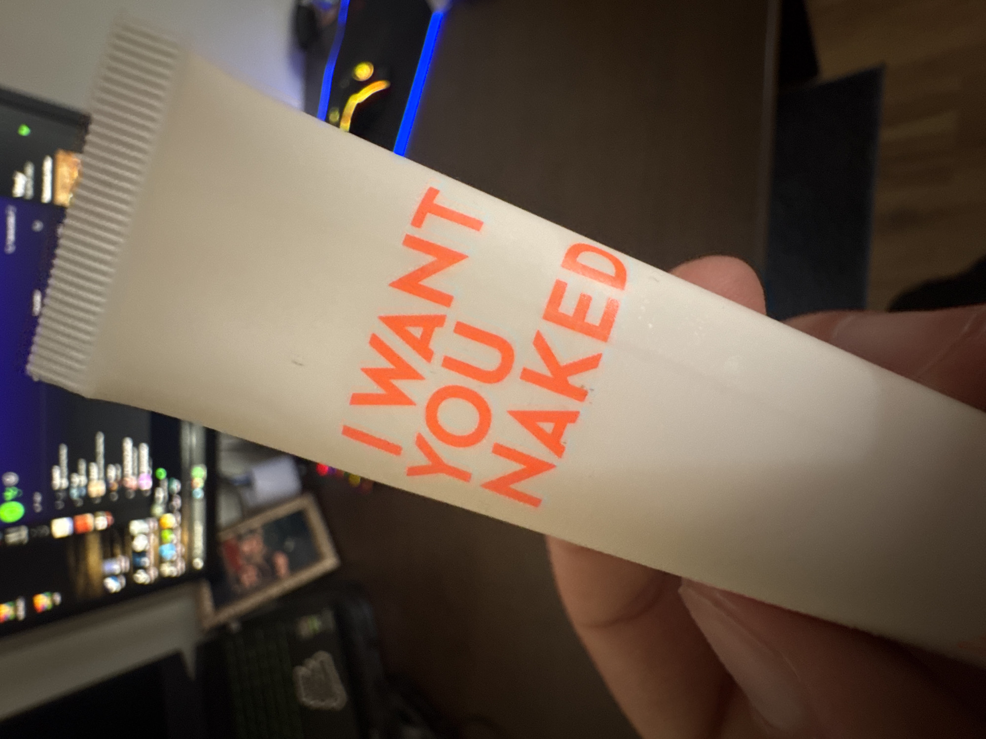 A hand holds a white tube with the words "I WANT YOU NAKED" printed in orange. The tube is slightly angled, with the top portion facing towards the upper left of the frame. The background includes a computer monitor displaying multiple icons and applications, along with a picture frame resting on a dark object. The surrounding environment consists of a dimly lit interior space with a dark, unidentifiable shape in the distance. The location, Varna, Bulgaria, cannot be determined from the image.