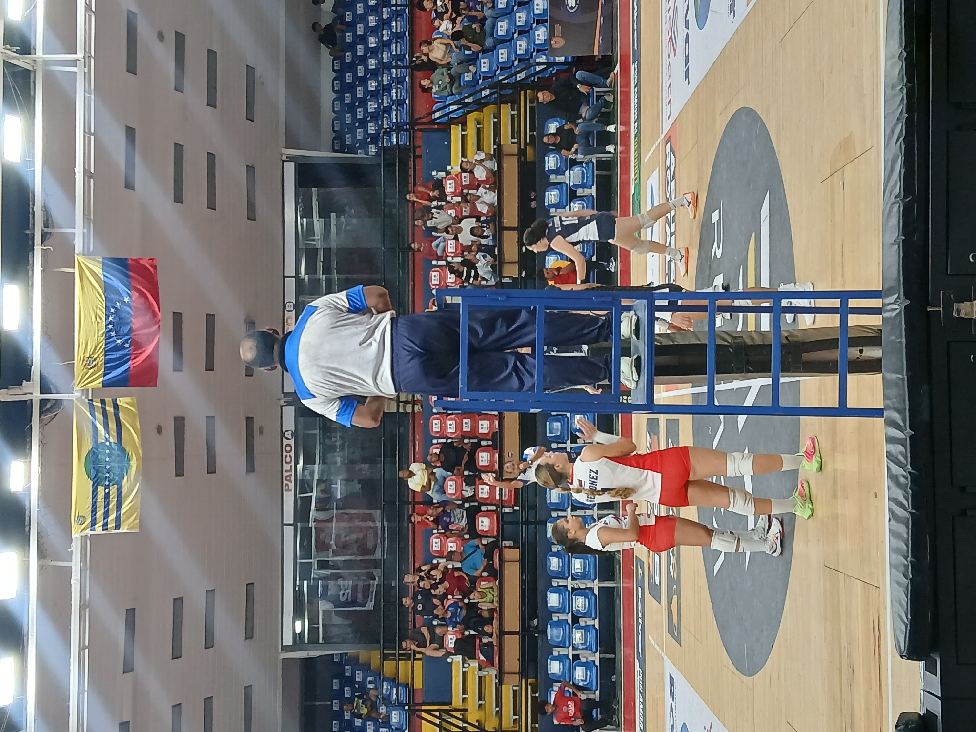 The image depicts a volleyball game indoors. The location is Ciudad Guayana, Venezuela, suggested by the Venezuelan flags hanging on the left. A referee stands on a blue platform. Two volleyball players wearing red and white uniforms are on the right side of the image. Another player in a dark uniform is preparing to hit the ball on the right side of the image near the center. Spectators fill the blue, yellow, and red seats in the background. The court has a large circular design in its center.