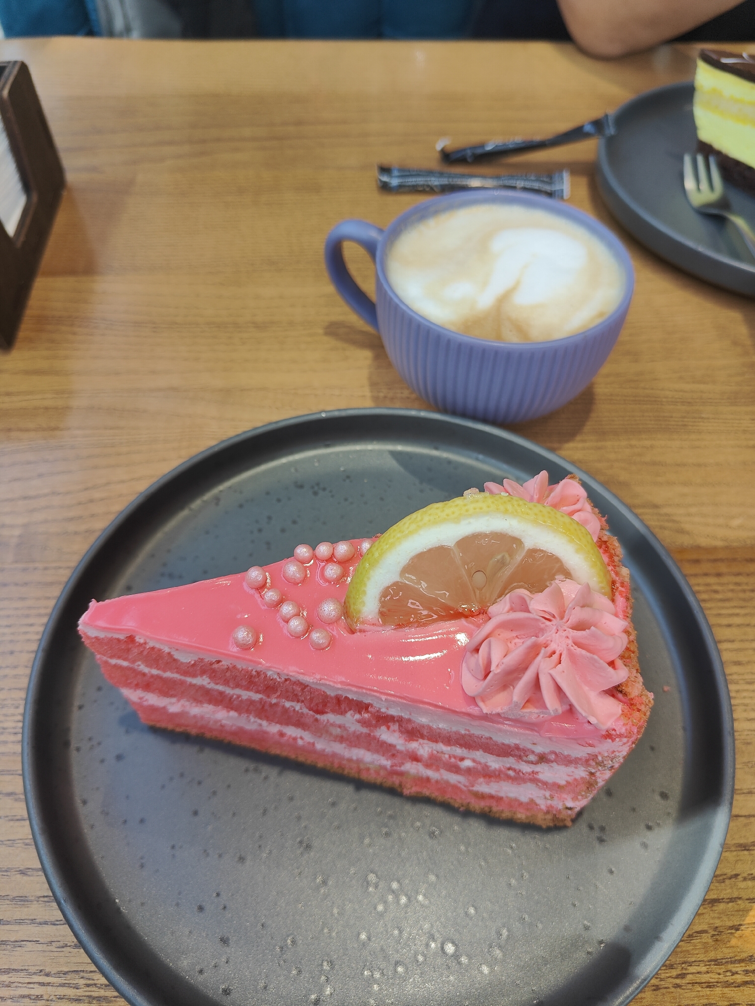This is a close-up, eye-level shot of a slice of pink layered cake with a slice of lemon and pink frosting on a dark grey plate, positioned on a light brown wooden table. To the left and slightly behind the cake, a purple mug filled with a latte sits on the table. In the background, another slice of cake, this one with yellow and white layers and a chocolate glaze, is on a similar dark grey plate with a fork. Two packets, possibly sugar or creamer, lie on the table between the mug and the second cake. The environment appears to be a cafe or restaurant in the city of Chornomorsk, Ukraine. The lighting is soft and natural, suggesting an indoor setting during the day. No individuals are clearly visible, but a hint of clothing in the background suggests at least one person is present.
