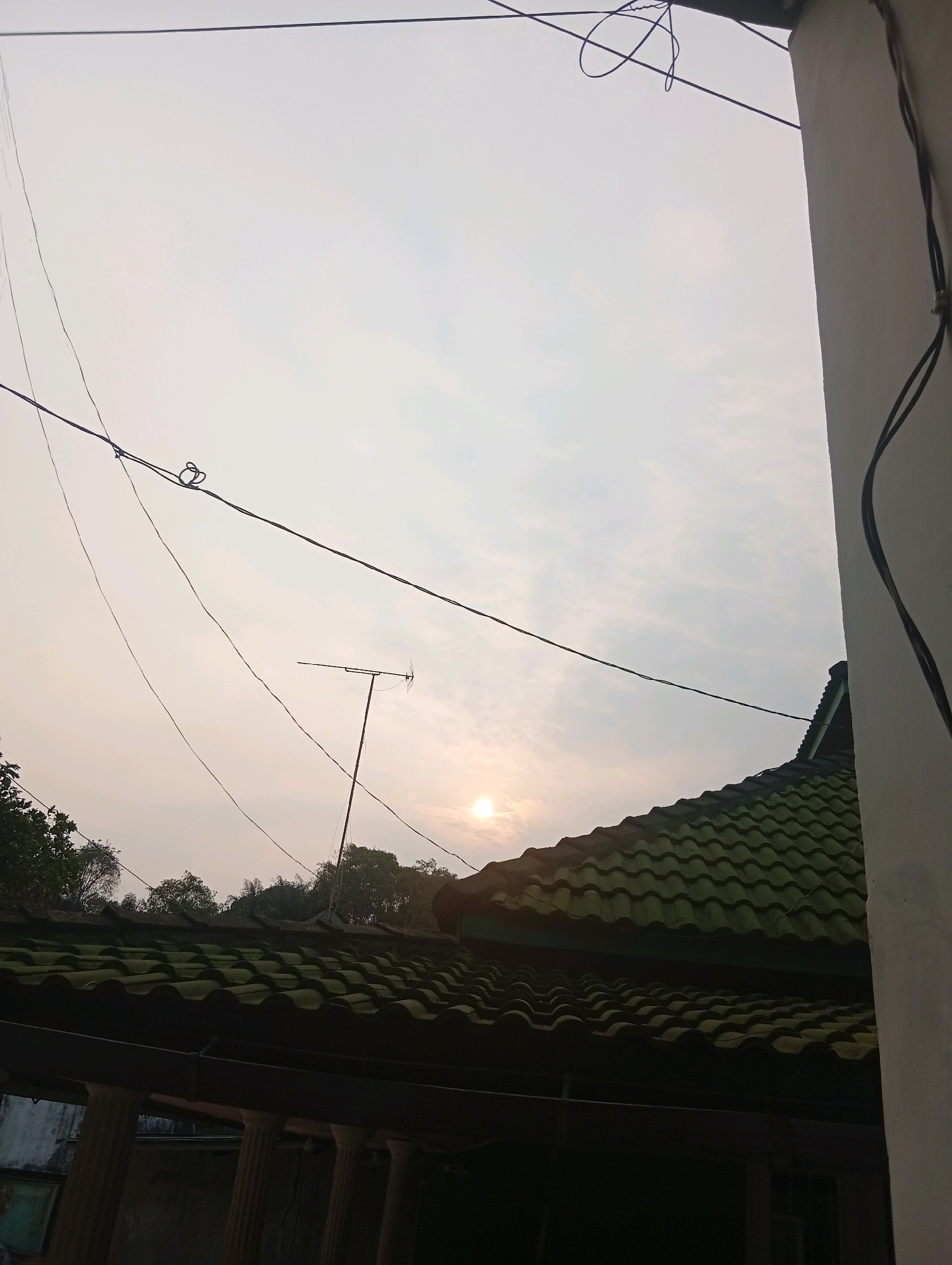 The image shows a view from a rooftop in Samarinda, Indonesia, looking upwards towards a hazy sky with the sun visible as a bright disc. Power lines crisscross the frame, and a television antenna is mounted on a pole. In the foreground, a portion of a green tiled roof is visible, with the underside of the roof structure and some pillars below. Trees can be seen in the background against the sky. The overall scene suggests an urban or semi-urban residential setting at dusk or dawn.
