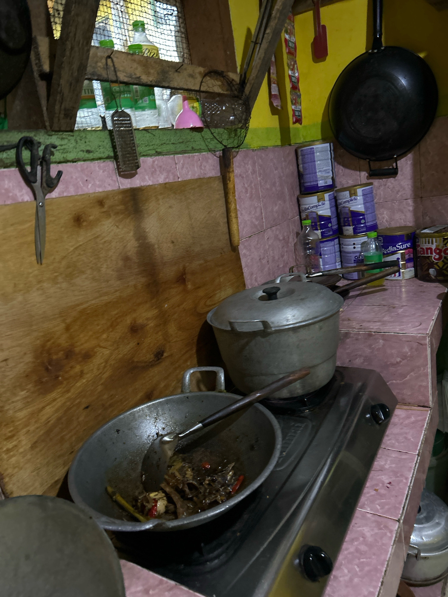 This is an indoor, medium-shot, eye-level, still photograph of a rustic kitchen counter and gas stove. The kitchen appears to be in Cihaurbeuti, Indonesia, given the context. The counter is covered in pink tile, and a wooden cutting board is leaning against the wall. On the stovetop, a wok containing food is being cooked, and a large pot sits next to it. Behind the stove, several cans of formula and other items are stacked on a shelf. A pair of scissors hangs from a hook on the wall. The overall impression is one of a functional, lived-in kitchen.