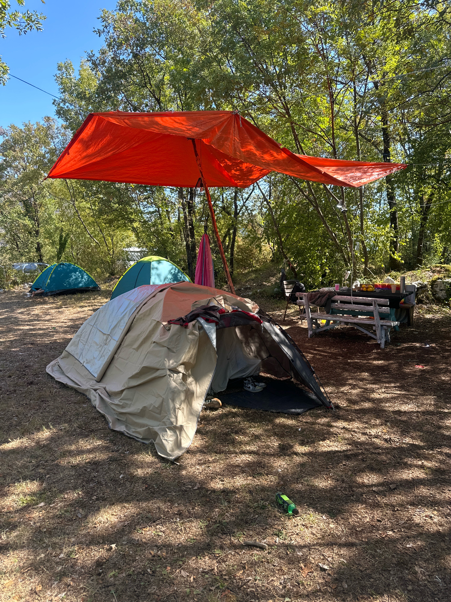 A campsite is set up in a wooded area with several tents. An orange tarp is stretched over a portion of the campsite, providing shade. A picnic table with benches is set up nearby, with food and drinks on the table. There is a green and yellow tent and a blue tent visible in the background, as well as a tan and brown tent in the foreground. The ground is covered in dry leaves and pine needles. The image was taken on a sunny day, with shadows cast by the trees. The location of this campsite is in the city of Cetinje, Montenegro.