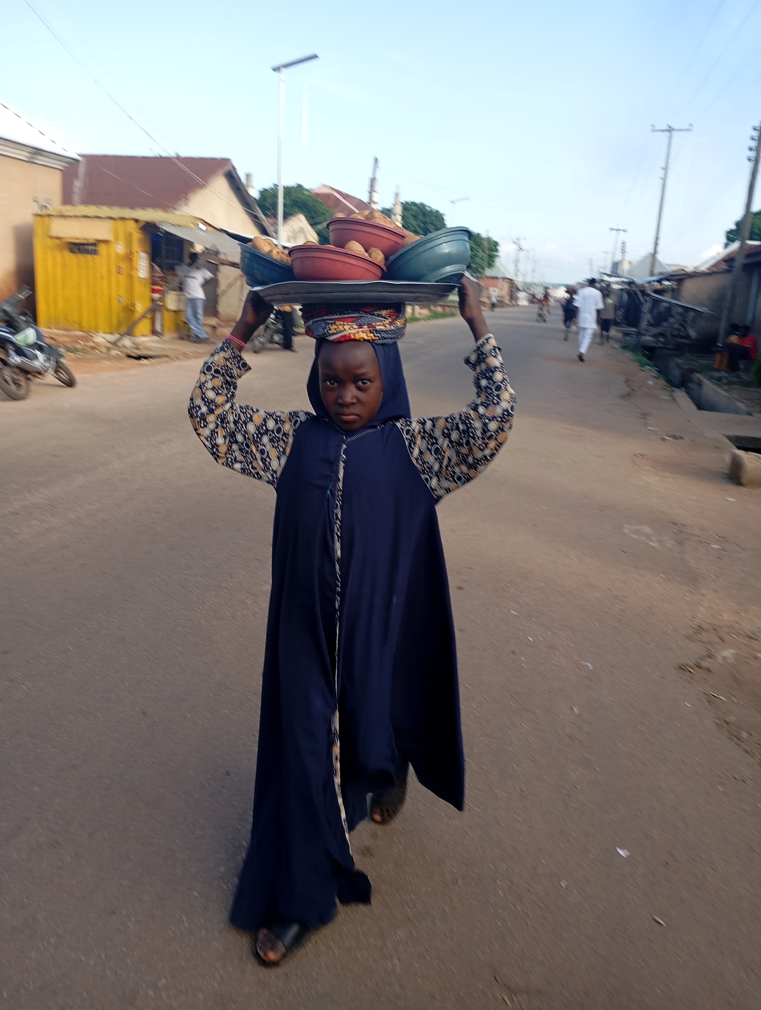 A young girl, likely of Fulani ethnicity, walks down a dusty street in Shendam, Nigeria, balancing a tray of food on her head. She is dressed in a long, dark blue garment and wears a patterned headscarf. The tray contains several bowls of food, including what appear to be potatoes and other vegetables. The street is lined with modest buildings, some with corrugated iron roofs, and a few motorcycles are parked along the side. In the background, other individuals can be seen walking and going about their day. The sky is clear and the lighting suggests it is late afternoon or early morning.