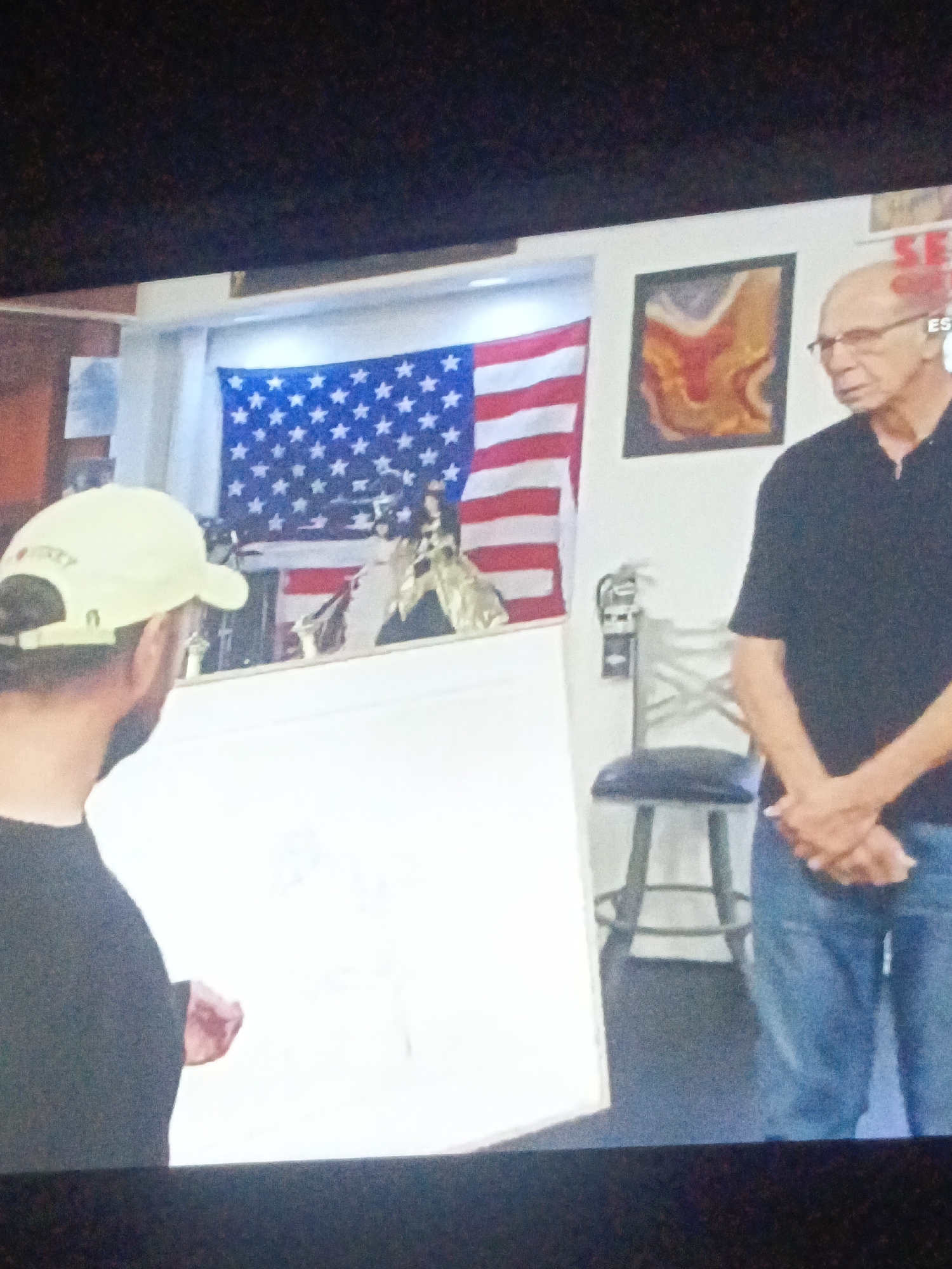 The image depicts an indoor scene featuring two men. One man, facing away from the camera, wears a green baseball cap and a black shirt. The other man, facing the camera, wears glasses, a black polo shirt, and blue jeans. He stands with his hands clasped. Behind them, a United States flag hangs above a display shelf with various objects. A piece of artwork hangs next to the flag, and a chair is positioned to the right of the man facing the camera. A fire extinguisher is also visible. The scene takes place indoors, though details that suggest a specific location within Juan Griego, Venezuela, are absent.