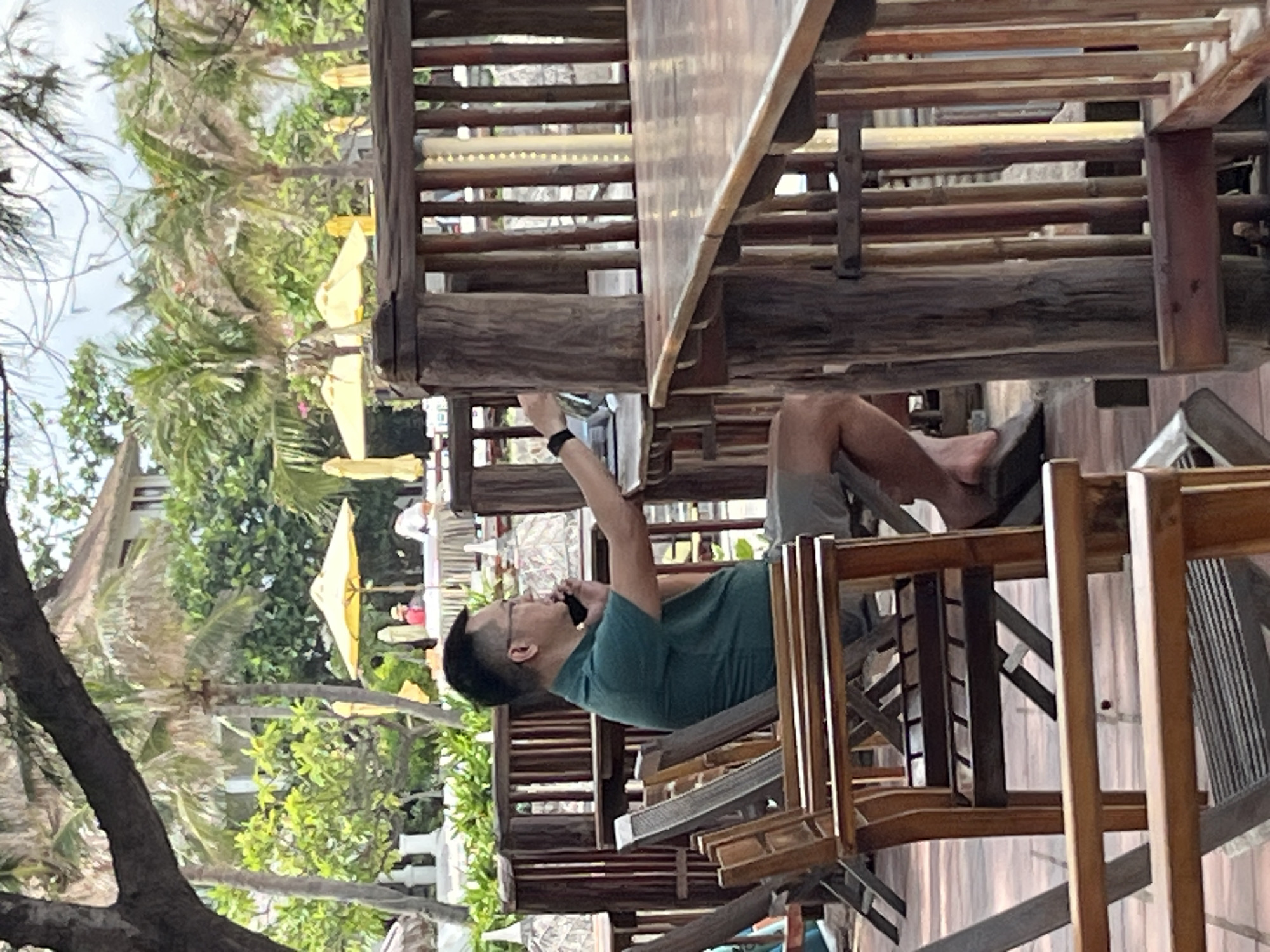 A man wearing glasses, a dark green polo shirt, and light-colored shorts sits in a chair at an outdoor table in Mũi Né, Vietnam. He is holding a phone to his left ear with his left hand and appears to be looking at a laptop or tablet on the table. His right arm is extended and his hand is near the device. The background shows palm trees, yellow umbrellas, and a building. The seating area features wooden chairs and tables, with a rustic fence made of bamboo and wood posts. The ground is tiled in a terracotta color.