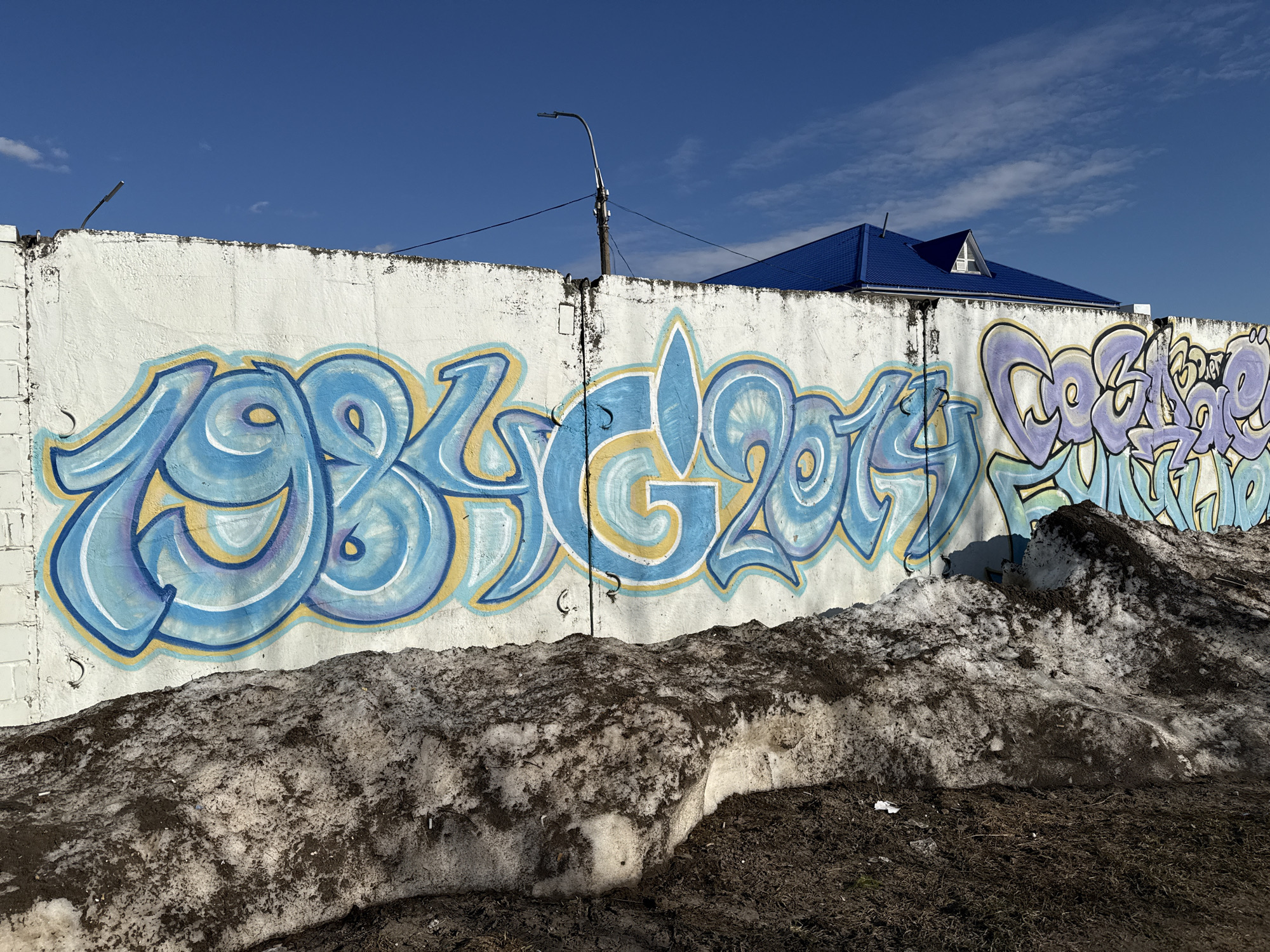 A white, segmented concrete wall is covered in graffiti of swirling blue, purple, and yellow paint that says "1984 Glory 2014" and other indiscernible words. The wall is in front of a pile of dirty snow, and behind the wall is a blue-roofed building and a utility pole. The sky is blue with some clouds. The location is in a city of Unknown, Unknown.