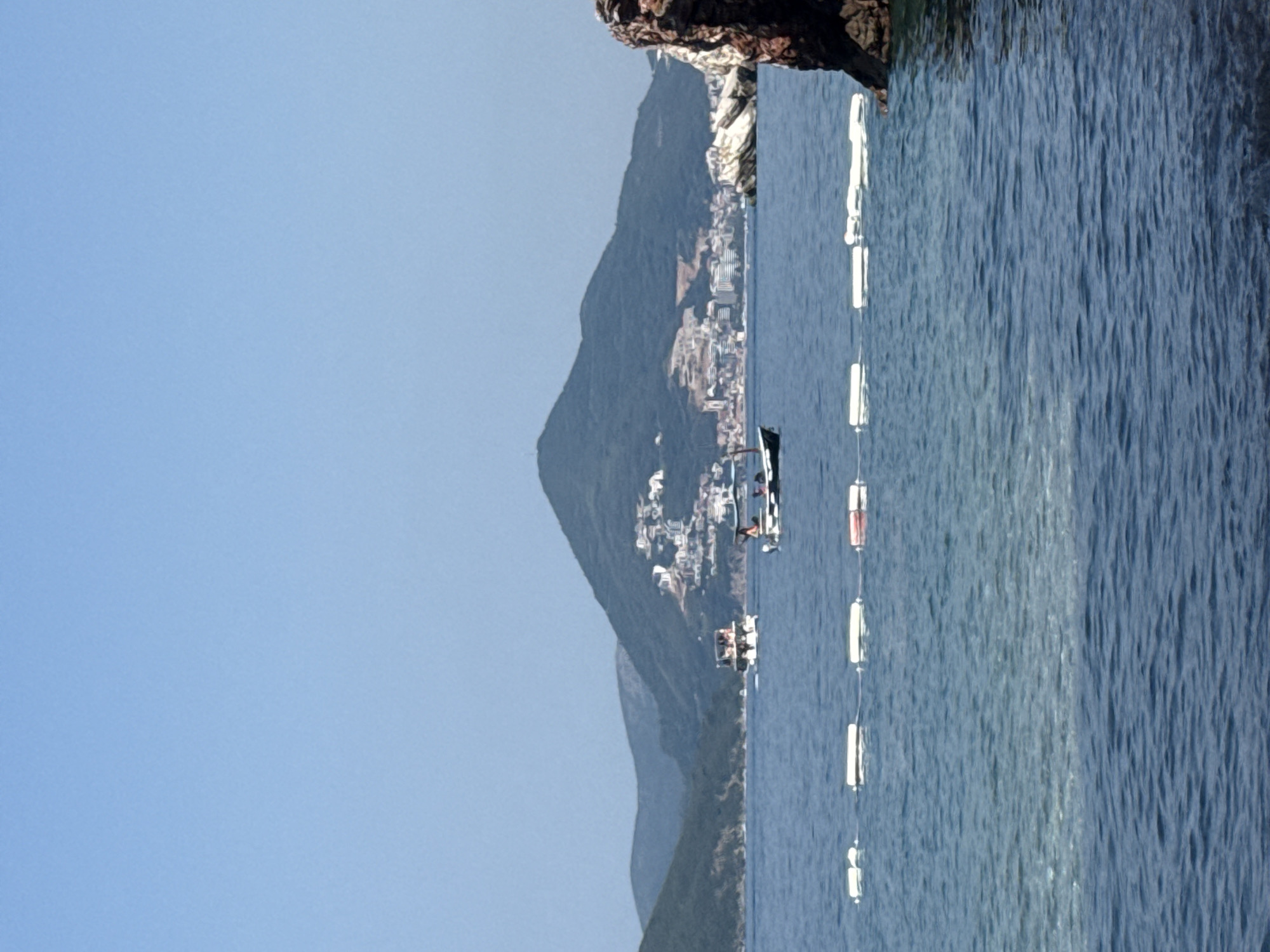 The image depicts a serene coastal scene with a clear blue sky and tranquil blue water. In the foreground, a rocky outcrop is visible on the left. The middle ground features a bay with several boats and a buoy line stretching across the water. The largest boat, a speed boat, is the most prominent, with people on board. Further in the background, a densely populated hillside town with buildings is nestled against lush green mountains, suggesting a Mediterranean or Adriatic coastal landscape. The overall impression is one of a bright, sunny day, likely in the late morning or early afternoon, given the quality of light. The scene evokes a sense of peaceful leisure and vacation. The location is identifiable as Boreti, Montenegro, based on the provided context. No visible text is present in the image.