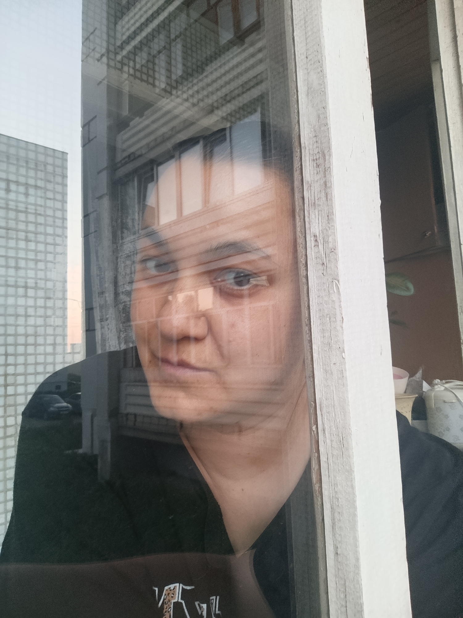 A person is looking out of a window. The window frame is painted white and appears old with chipped paint. Through the window, a building with a grid-like facade can be seen, and the reflection of another building with horizontal architectural elements is visible in the glass. The environment outside the window is urban, with a car partially visible. Inside the room, other objects are visible in the background. Based on the details and context, the location is Chaikovsky, Russia.