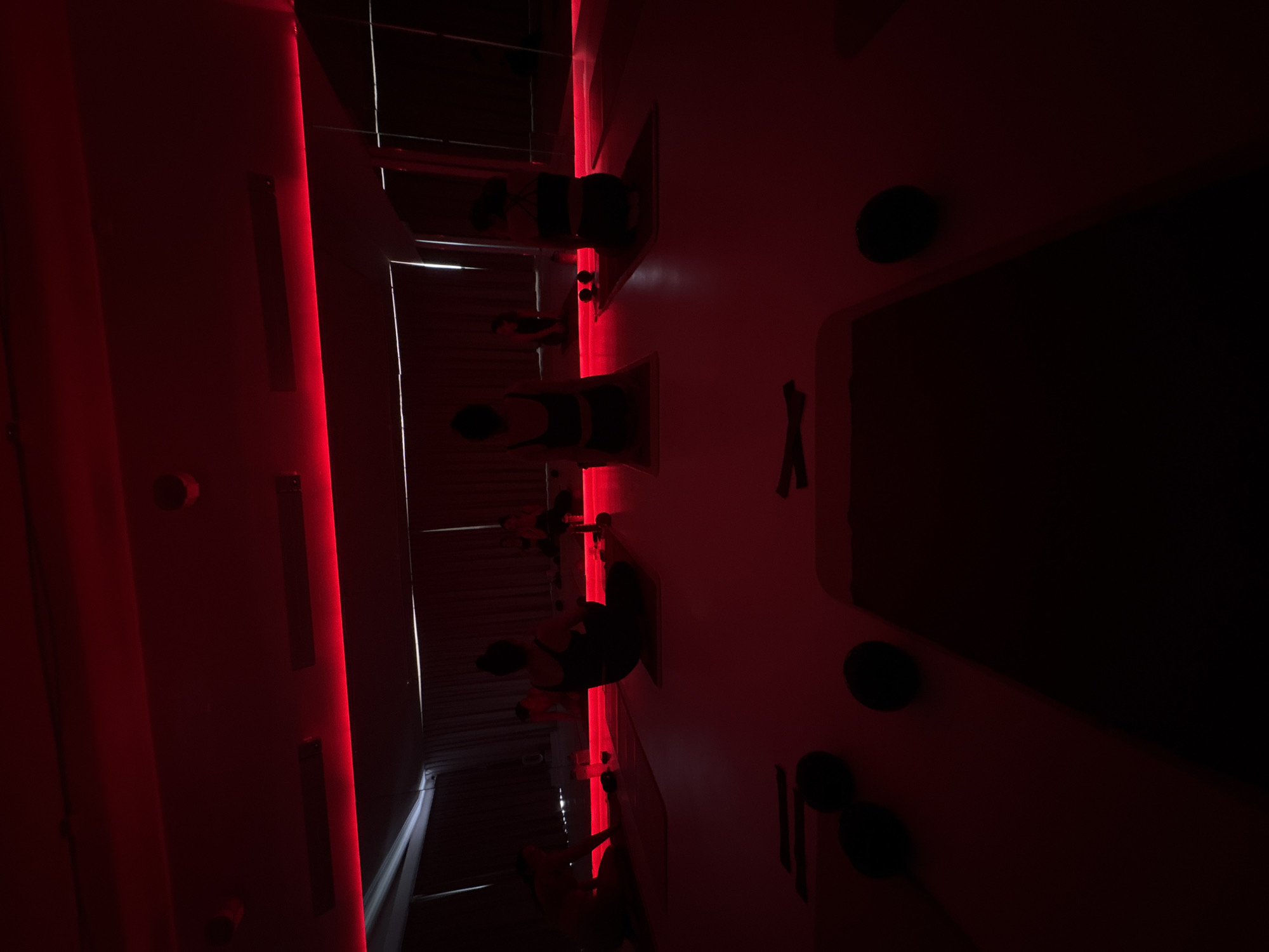 The scene depicts an interior view of a fitness or wellness studio in Mexico City, Mexico, illuminated predominantly by a deep red light. Multiple individuals, primarily visible from behind, are seated or kneeling on individual dark exercise mats on a light-colored floor. A large mirrored wall at the back reflects the room and some of the participants. Red LED strip lighting runs along the top edge of this mirrored wall, casting a crimson hue throughout the space.
In the foreground, a dark exercise mat is partially visible, with several small, dark, spherical objects and what appear to be exercise bands or straps placed near it. Further back, several individuals are positioned on their mats. One person in a dark tank top and leggings is seated facing away from the camera in the center. To their left, another individual is similarly seated. To their right, a third person is seated on a mat with a clear bottle and small dark spherical objects nearby. In the reflection of the mirrored wall, additional individuals are visible, also seated or kneeling on mats. The environment is orderly, suggesting a structured class or session. The ceiling features recessed lighting fixtures and what appear to be speakers or ventilation grills. Dark curtains are visible behind the mirrored wall in the background.