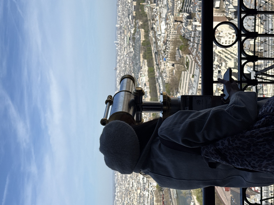 The image captures an individual, seen from behind, positioned on an elevated observation deck in Paris, France. The subject, appearing female, wears a grey beret, a grey coat, and a patterned backpack on her left shoulder. She is actively looking through the eyepiece of a silver-colored, coin-operated viewing telescope, which is mounted on a dark stand with a gold-colored base. Her right hand holds a dark-colored smartphone or similar device near the telescope's control panel or coin slot. A decorative black metal railing with circular and scrolled elements is visible in the immediate foreground. Beyond the railing, a panoramic daytime cityscape of Paris unfolds under a clear blue sky with scattered white clouds. The view includes dense urban architecture, green areas, the winding Seine River in the mid-left, and a cluster of distant modern skyscrapers on the horizon. The lighting indicates the sun is behind the observer, silhouetting the individual and the telescope.
