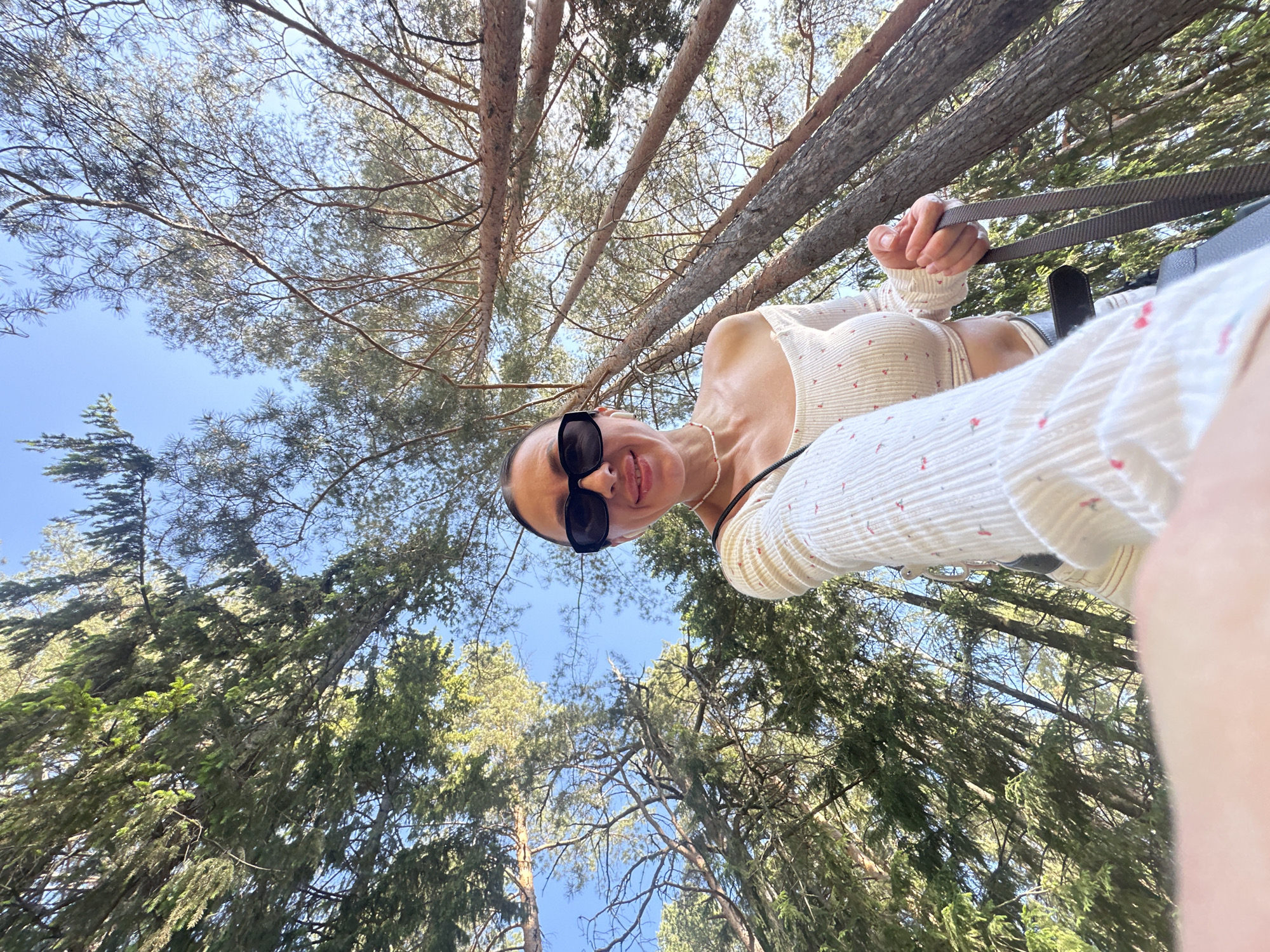 The photograph is taken from a low angle, looking upwards into a dense forest canopy. The sky is visible through the branches, indicating an outdoor setting. A woman wearing sunglasses and a patterned top is looking down at the camera. She is holding onto a gray strap. The location appears to be a wooded area, potentially within the city of Votkinsk, Russia, or in its vicinity.