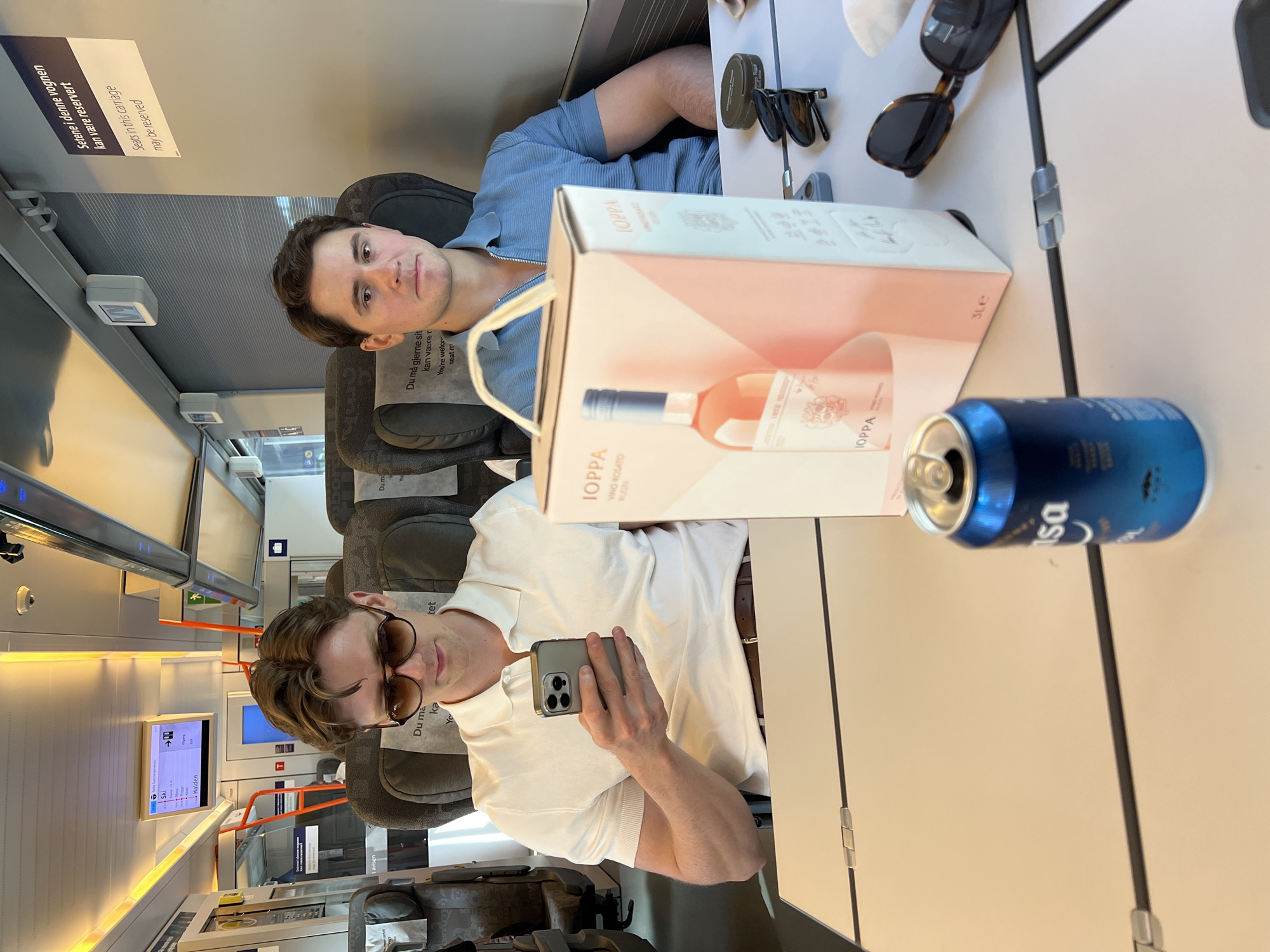 Two men are seated on a train. The man on the left is wearing a white polo shirt and sunglasses, and is holding a smartphone. The man on the right is wearing a blue polo shirt. In front of them is a table with a box of wine, a can of soda, and sunglasses. The train appears to be moving.