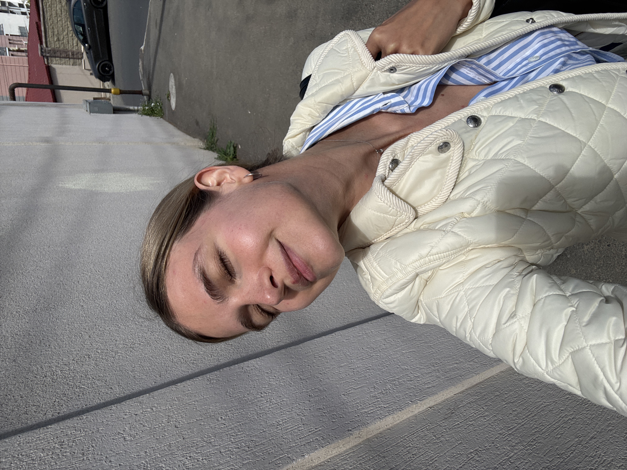 A close-up, rotated photo shows a person, likely female, from the chest up, with her head tilted. She has light brown hair styled back, closed eyes, and a soft, subtle smile, conveying a peaceful or serene expression. She is wearing a cream or off-white quilted jacket with snap buttons, revealing a blue and white striped shirt underneath. A delicate silver-colored chain necklace with a small pendant is visible around her neck, and a small silver hoop earring is in her left ear. One hand is partially visible inside her jacket, near the collar.
The background, due to the image's 90-degree counter-clockwise rotation, appears as a grey, textured surface, likely asphalt or concrete ground when viewed upright. In the top left corner (when upright), a black car is partially visible, along with parts of a building or structure and a yellow-tipped metal pipe railing. Small patches of green vegetation are growing along the edges of the paved area. Bright sunlight is evident, creating clear shadows, indicating it is daytime. No visible text is present in the image.