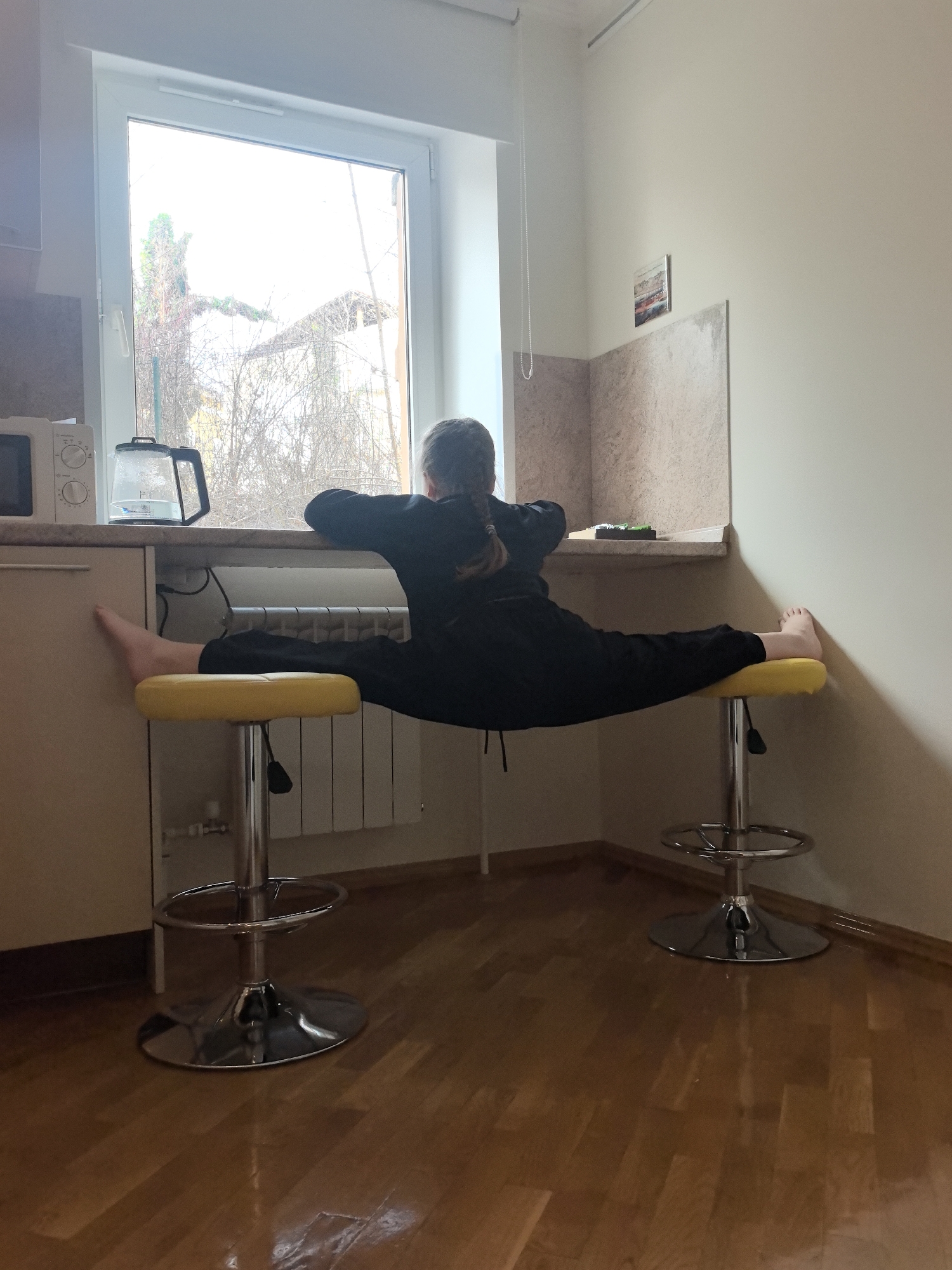 A young person with braided hair, wearing a dark-colored jumpsuit, is performing a split on two yellow bar stools in what appears to be a kitchen. The person's torso is resting on a counter beneath a window that reveals a view of bare trees, potentially indicating a cold season. In the kitchen, there are standard appliances such as a microwave and a kettle. The floor is made of wooden parquet. A radiator is visible behind one of the stools. A picture is affixed to the wall. Based on the overall aesthetic, this could be a residential interior in Nizhny Novgorod, Russia.