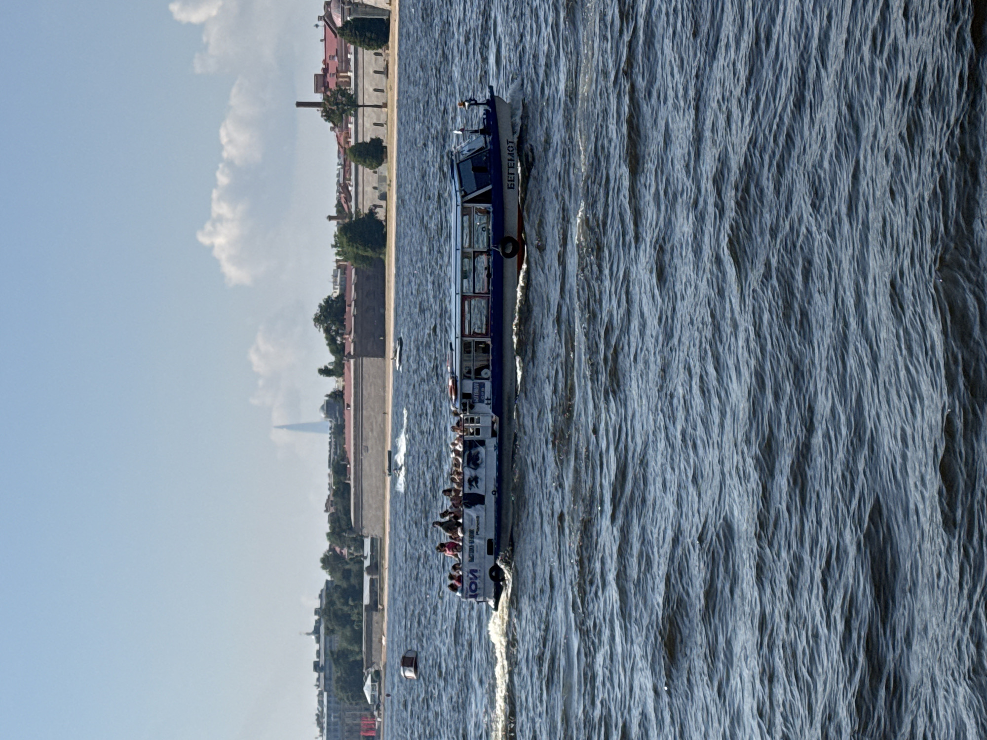 The image depicts a passenger boat named "BEGEMOT" cruising on a body of water, likely the Neva River in Saint Petersburg, Russia. The boat is filled with passengers, many standing and observing the surroundings. The boat has a blue and white hull with windows along its side. In the background, the cityscape of Saint Petersburg is visible, characterized by historical buildings with red tiled roofs and a distinctive tall, slender spire in the distance, possibly the Lakhta Center. The water is choppy, indicating a breezy day. The sky is clear with a few scattered clouds.