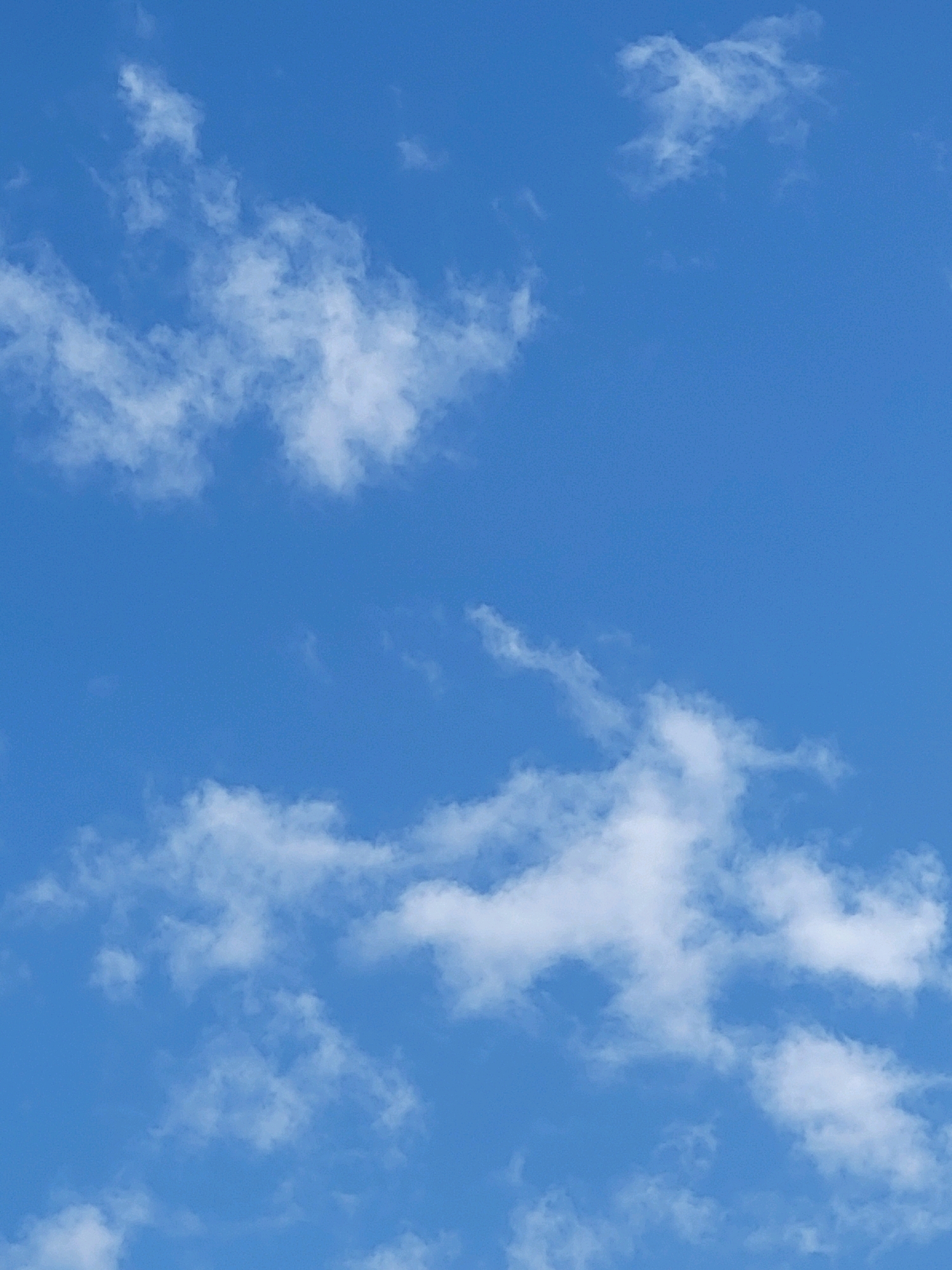 The image is a close-up, high-angle shot of a bright blue sky with scattered white cumulus clouds. The clouds are wispy and varied in density, with some appearing more solid and others more ethereal. The lighting suggests it is daytime, with the sun likely positioned high in the sky, casting a clear light on the clouds. There are no discernible landmarks or environmental features visible that would suggest a specific location within the city of Jelgava, Latvia. The image exclusively depicts the sky and clouds.