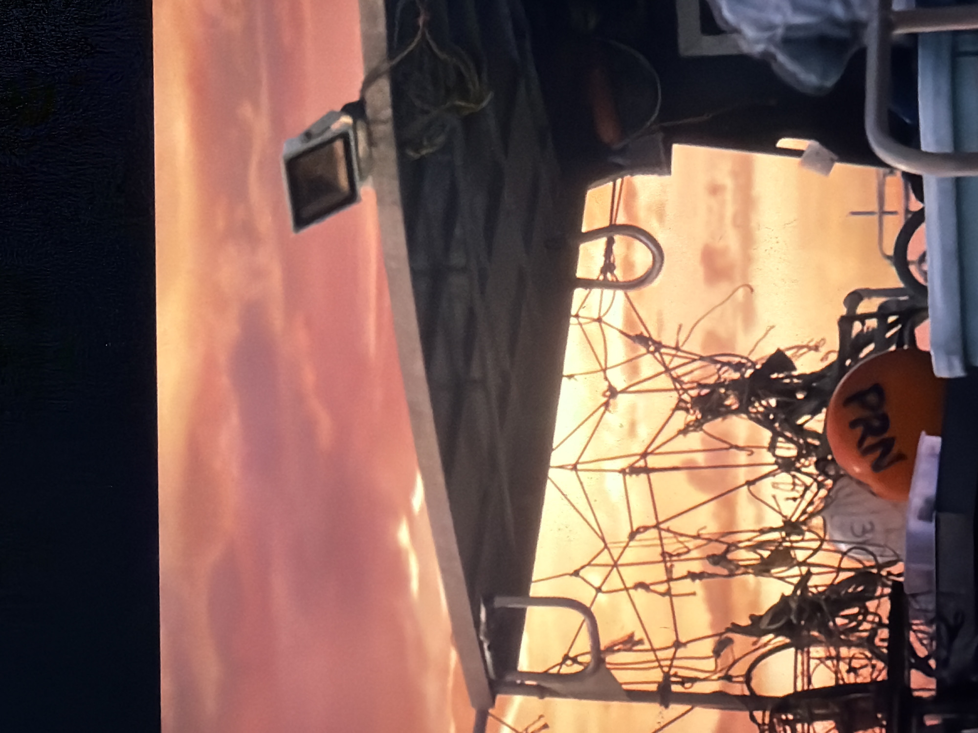 The image shows a close-up, angled view of the deck of a boat at sunset. The sky is a vibrant mix of orange, pink, and red clouds. In the foreground, there is a structure with a dark-colored roof supported by metal poles. Hanging from the underside of the roof are metal rings and a complex network of ropes and rigging. On the deck, an orange buoy with black lettering is visible, appearing to spell out "PRN". A white garment with the number "3" on it is also partially visible. To the right, a dark structure, possibly part of the boat's cabin, is present, with a person's arm and shoulder visible wearing a grey garment. The scene is consistent with a fishing boat or similar vessel operating at sea, with the sunset creating dramatic lighting. The location in Georgioupoli, Greece, is not discernible from the visual elements within the image itself, as there are no identifiable landmarks, signage, or architectural features specific to that city.