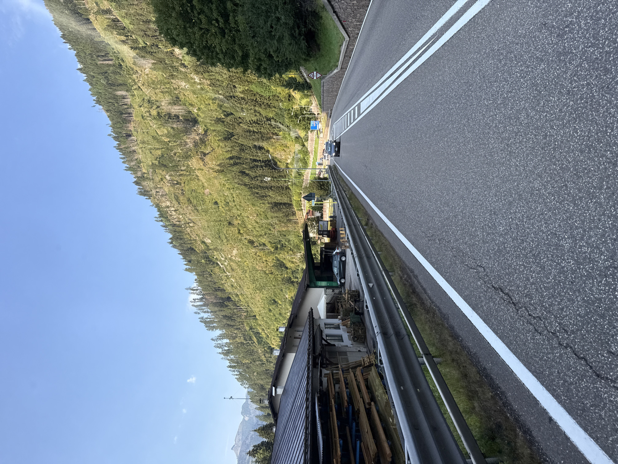 The image shows a paved road with double white lines in the foreground, indicating a two-lane highway. The road is flanked by a metal guardrail on the left and a stone-lined embankment on the right. On the left side of the road, there is a building with a dark, sloped roof, and a stack of lumber is visible next to it. Further down the road, two cars are driving, and the road curves to the right, leading into a mountainous area covered in pine trees. The sky is clear and blue. Based on the context provided by other images in the set, this scene is located in Predazzo, Italy.