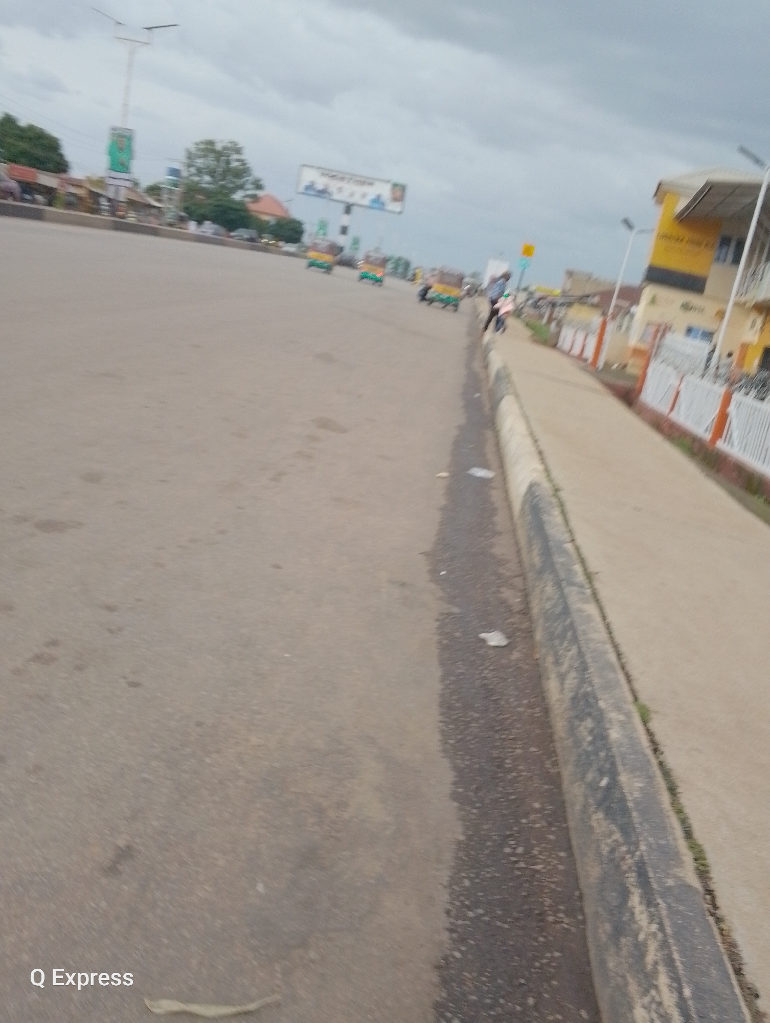 The media file depicts a street scene in Jos, Nigeria. The foreground is dominated by a wide asphalt road, with a concrete curb and sidewalk running along the right side. The road appears to be wet in patches, suggesting recent rain. There are a few scattered pieces of litter on the road and sidewalk.
In the middle ground, several three-wheeled vehicles, commonly known as "keke napeps" or auto-rickshaws, are visible. Some appear to be moving on the road. Beyond the vehicles, there are buildings lining the street, some of which are brightly colored, including a prominent yellow building with an awning. A white fence runs along the sidewalk in front of some of these buildings.
In the background, there are trees, utility poles, and billboards. One billboard displays images of people, though the text is largely illegible due to the image quality and distance. The sky is overcast, indicating cloudy weather, and the overall lighting suggests it might be late afternoon or early evening.
The scene appears to be a typical urban or semi-urban environment in Nigeria, likely a commercial or residential area with ongoing street activity. There are no discernible emotions conveyed by any individuals, as they are too small and indistinct to observe. The visible text in the image is "Q Express" in the bottom left corner.