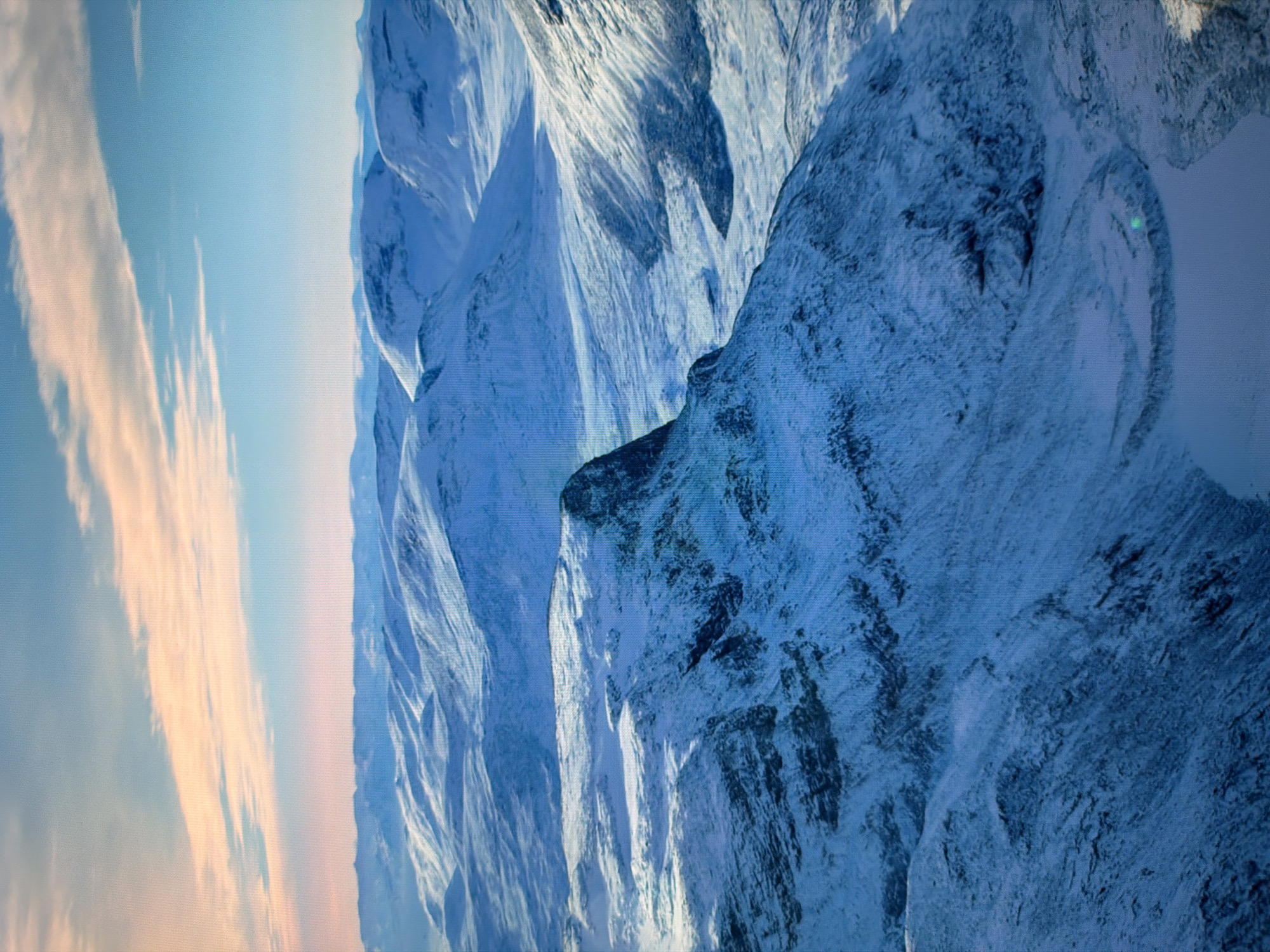 The image displays a vast, snow-covered mountainous landscape under a pale, cloud-streaked sky. The terrain is characterized by rolling hills and sharp peaks, all blanketed in white snow, suggesting a winter or early spring environment. Shadows are cast across the snow-covered surfaces, highlighting the contours and textures of the land. The sky transitions from a light blue near the horizon to a deeper blue overhead, with wispy, pale pink and orange clouds near the top of the frame. No human presence or artificial structures are visible. The scene is entirely natural and appears to be a remote, undeveloped area. This image is not from Kyiv, Ukraine, as the landscape is mountainous and snow-covered, unlike the predominantly flat terrain of Kyiv.