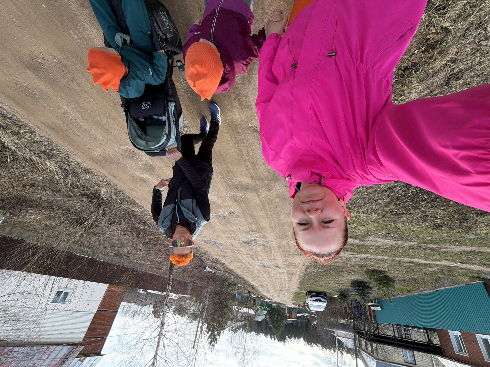 A woman in a pink jacket takes a selfie of herself, a woman in sunglasses and a gray/black jacket, and two children with orange hats walking on a dirt road in Chaikovsky, Russia. One child is in a stroller. Buildings are on either side of the road. A car is parked further up the road. The sky is overcast. The trees are mostly bare. The ground has dry vegetation.