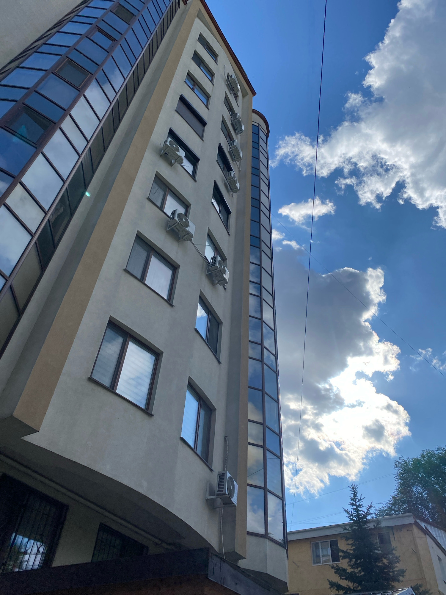 This is a low-angle, full-shot photograph of a modern, multi-story building in Chișinău, Moldova, under a partly cloudy blue sky. The building is primarily light beige with a rounded section covered in dark, reflective glass windows that mirror the sky and clouds. The other facade of the building features numerous windows and several outdoor air conditioning units mounted on the exterior walls. The windows are rectangular with dark frames, and some reflect the sky, while others show blinds or interior views. The building appears to be residential or commercial.
In the background, a smaller, yellow building with a tiled roof is partially visible, with a small evergreen tree in front of it. Overhead, two dark electrical wires stretch across the frame, intersecting the sky. The overall impression is one of urban architecture on a bright, possibly warm, day. No people or animals are visible in the frame. The street level is not clearly depicted, but some barred windows suggest a basement or ground-floor access.