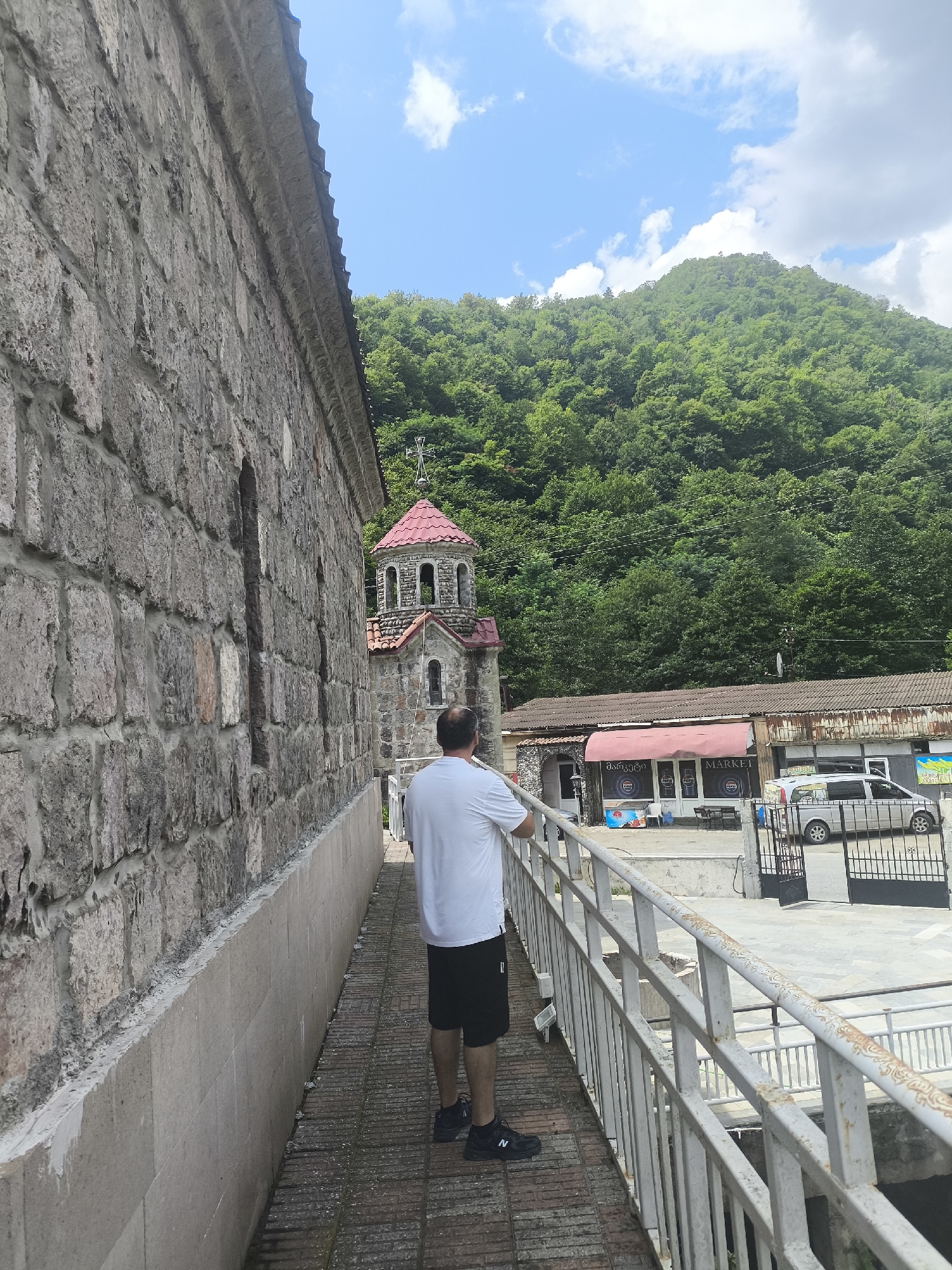 A man in a white t-shirt and black shorts stands on a walkway next to a stone building. He is facing away from the camera, looking towards a small church with a red roof and a steeple. In the background, there are trees on a hillside and a building that appears to be a market. The sky is blue with some clouds. The scene is located in Zvare, Georgia.